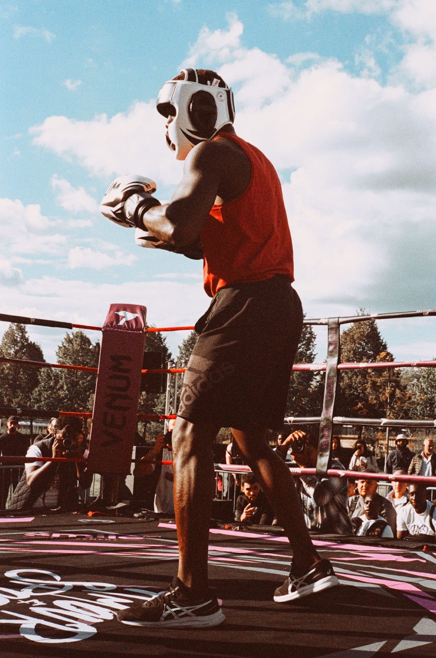 Boxeur en débardeur rouge en garde près du coin, photographes et public alignés derrière les cordes.