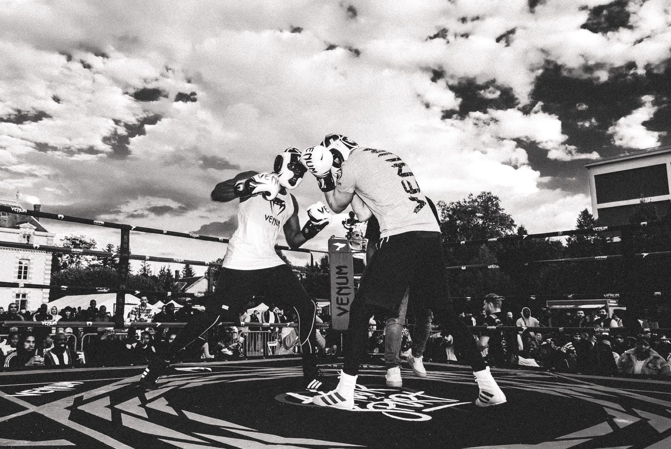 Noir et blanc : deux boxeurs au corps à corps au centre du ring, sous un ciel de gros nuages, stands en arrière-plan.