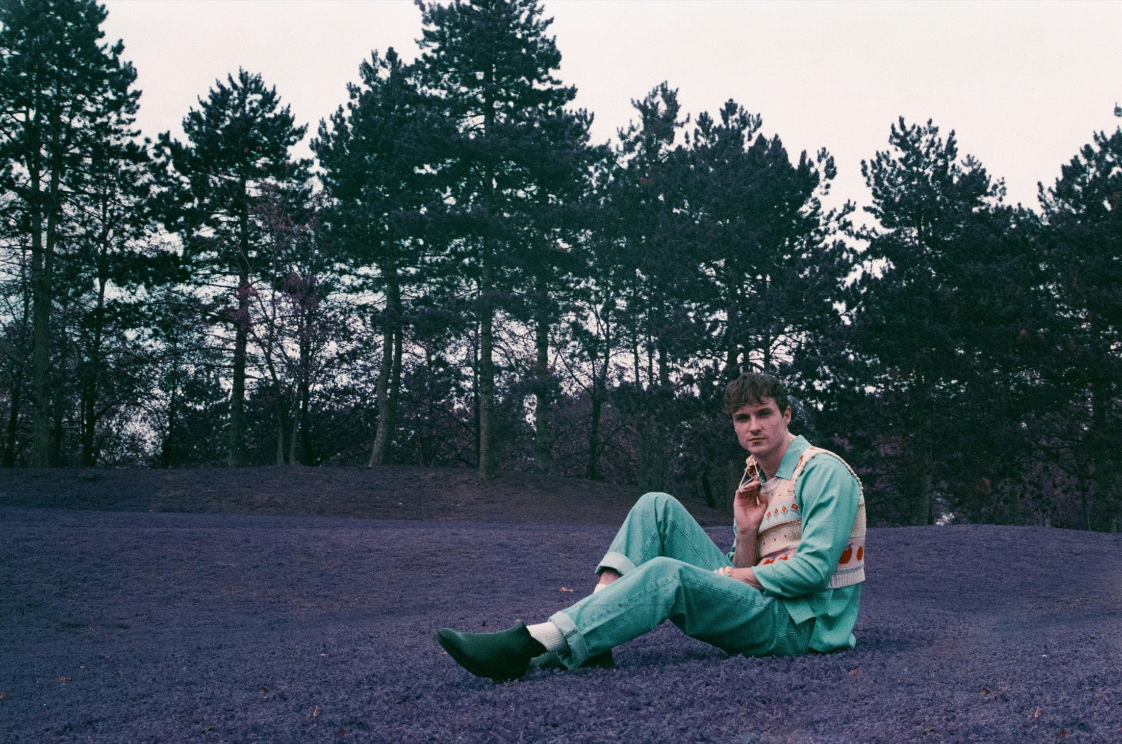 Homme assis sur l'herbe en denim vert d'eau et gilet à motifs, la pelouse virée au violet par le film, des pins derrière.