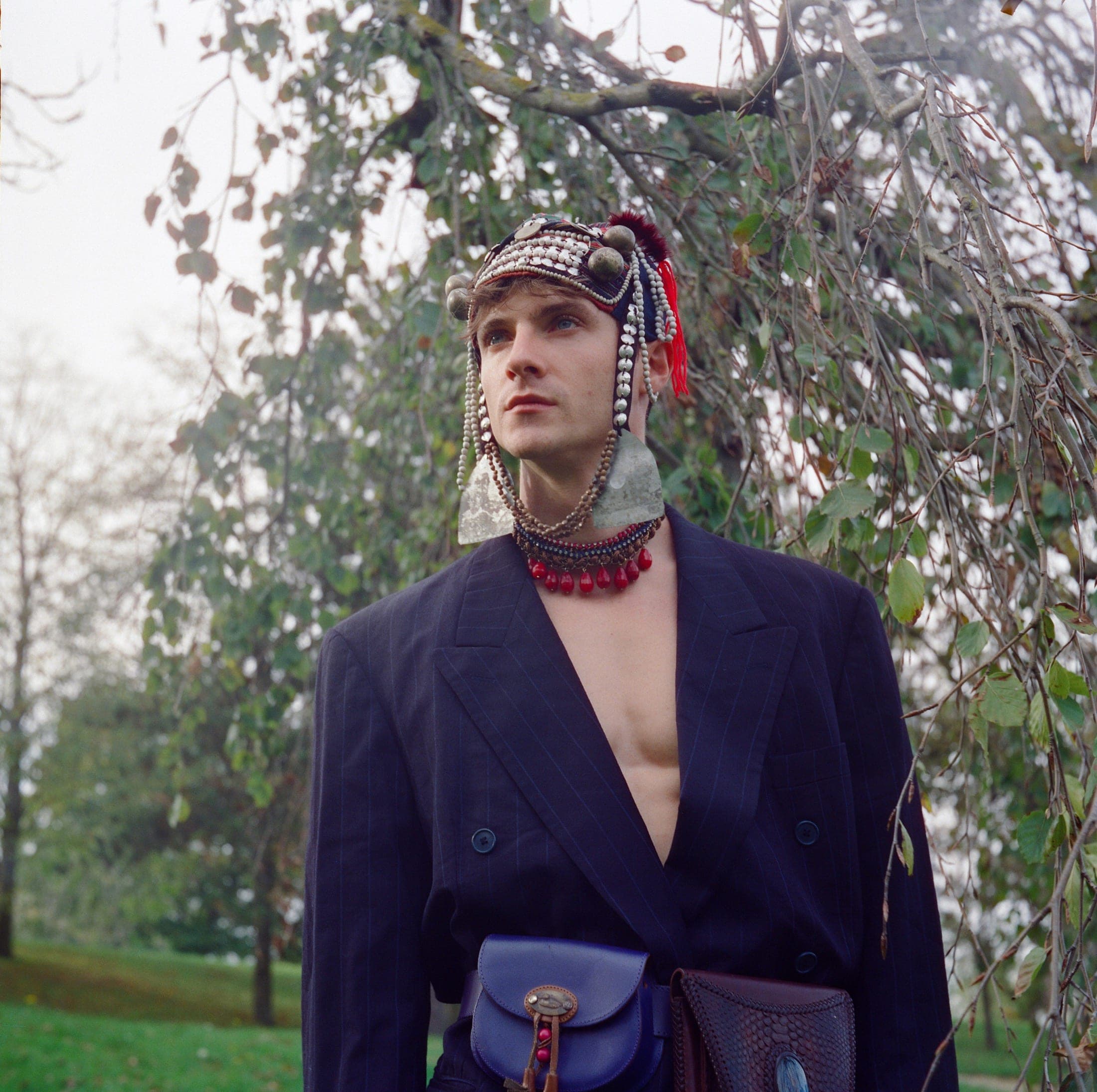 Sous un arbre pleureur, coiffe brodée de perles à pompon rouge, veste à fines rayures portée à même la peau et collier de perles rouges.