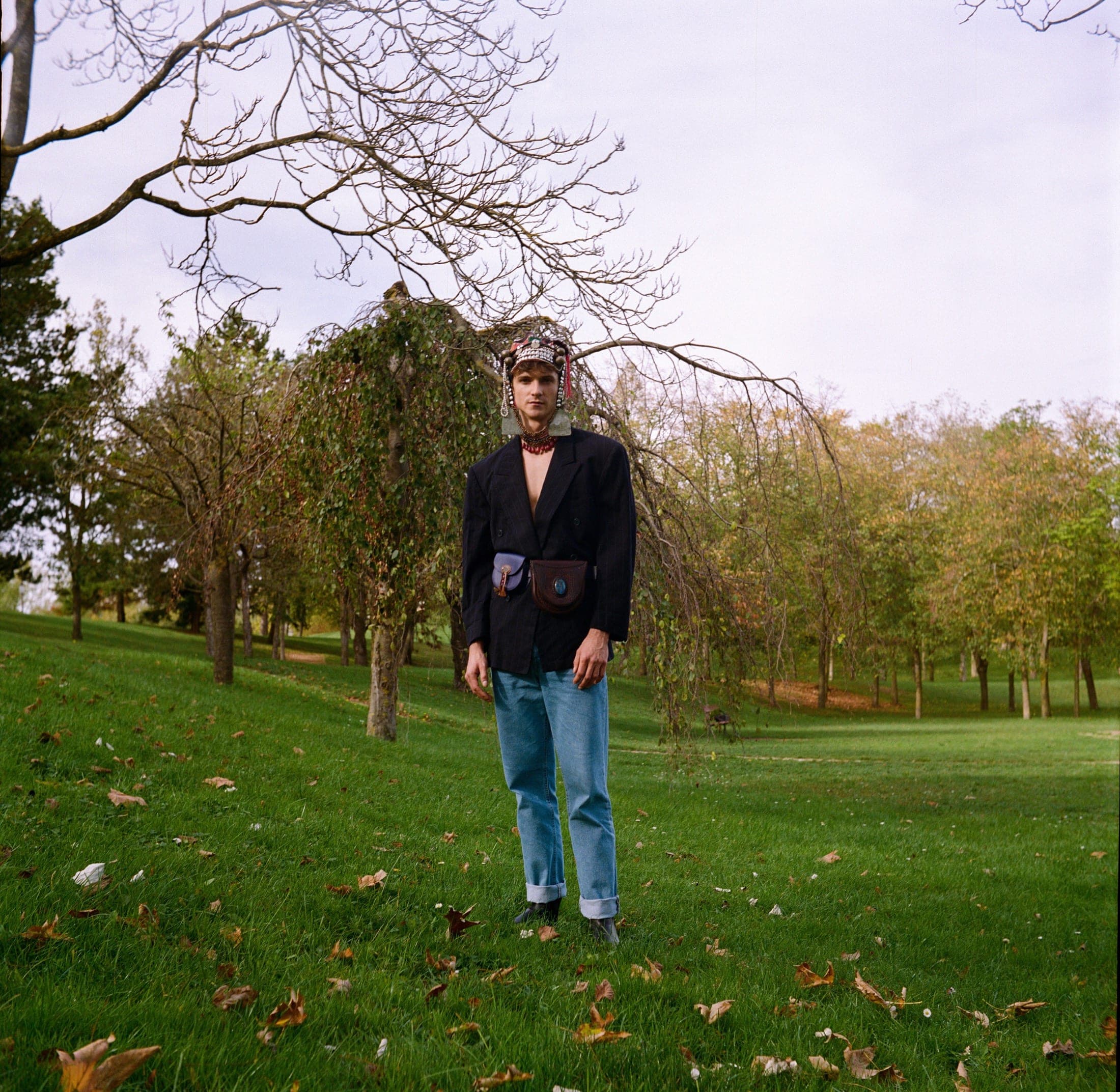 Au milieu d'une pelouse jonchée de feuilles, coiffe perlée, veste sombre ouverte et jean clair, arbres d'automne en fond.