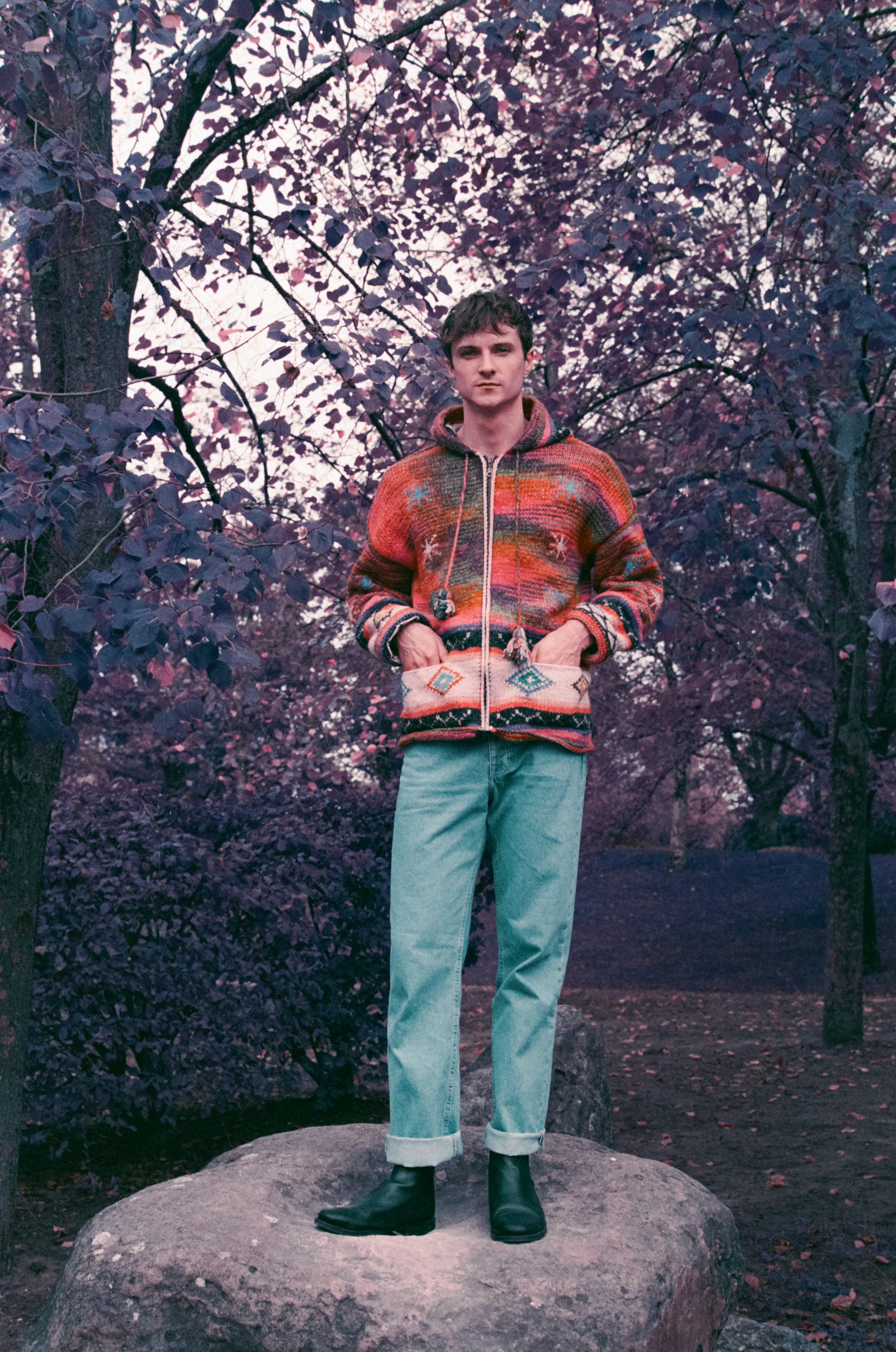 Debout sur un rocher, un homme en gilet zippé tricoté orange et turquoise, jean clair retroussé sur des bottines noires. Au-dessus, le feuillage a viré au violet.