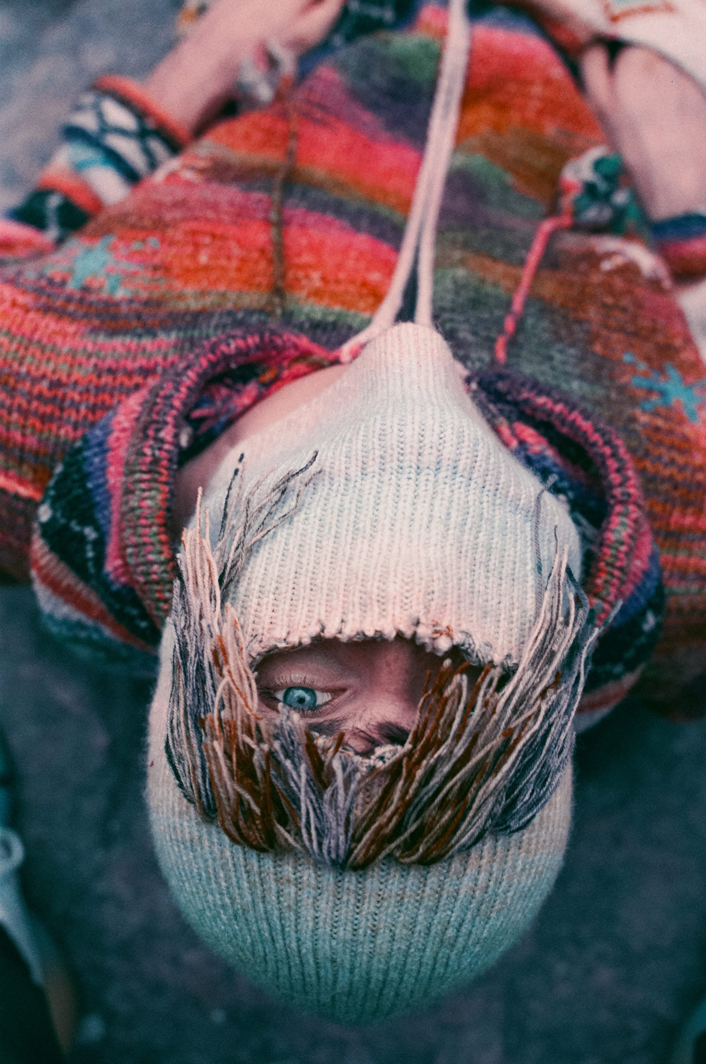 La tête renversée vers l'objectif, un œil bleu dans la fente d'une cagoule écrue frangée de laine, le col du gilet multicolore autour du visage.