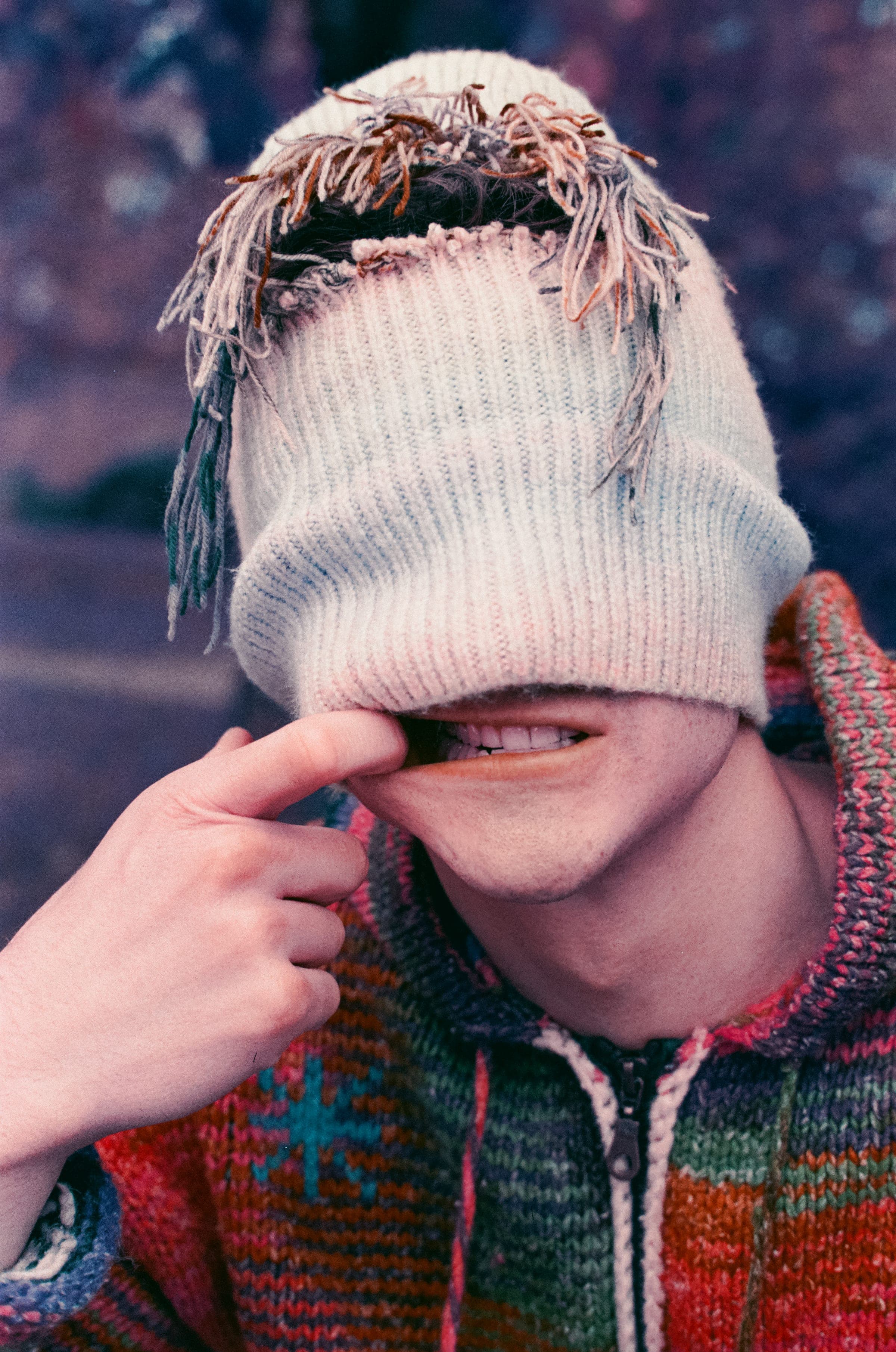 Cagoule écrue rabattue sur les yeux, franges de laine en bataille. Un doigt étire le coin de la bouche en sourire, au-dessus du col du gilet tricoté.