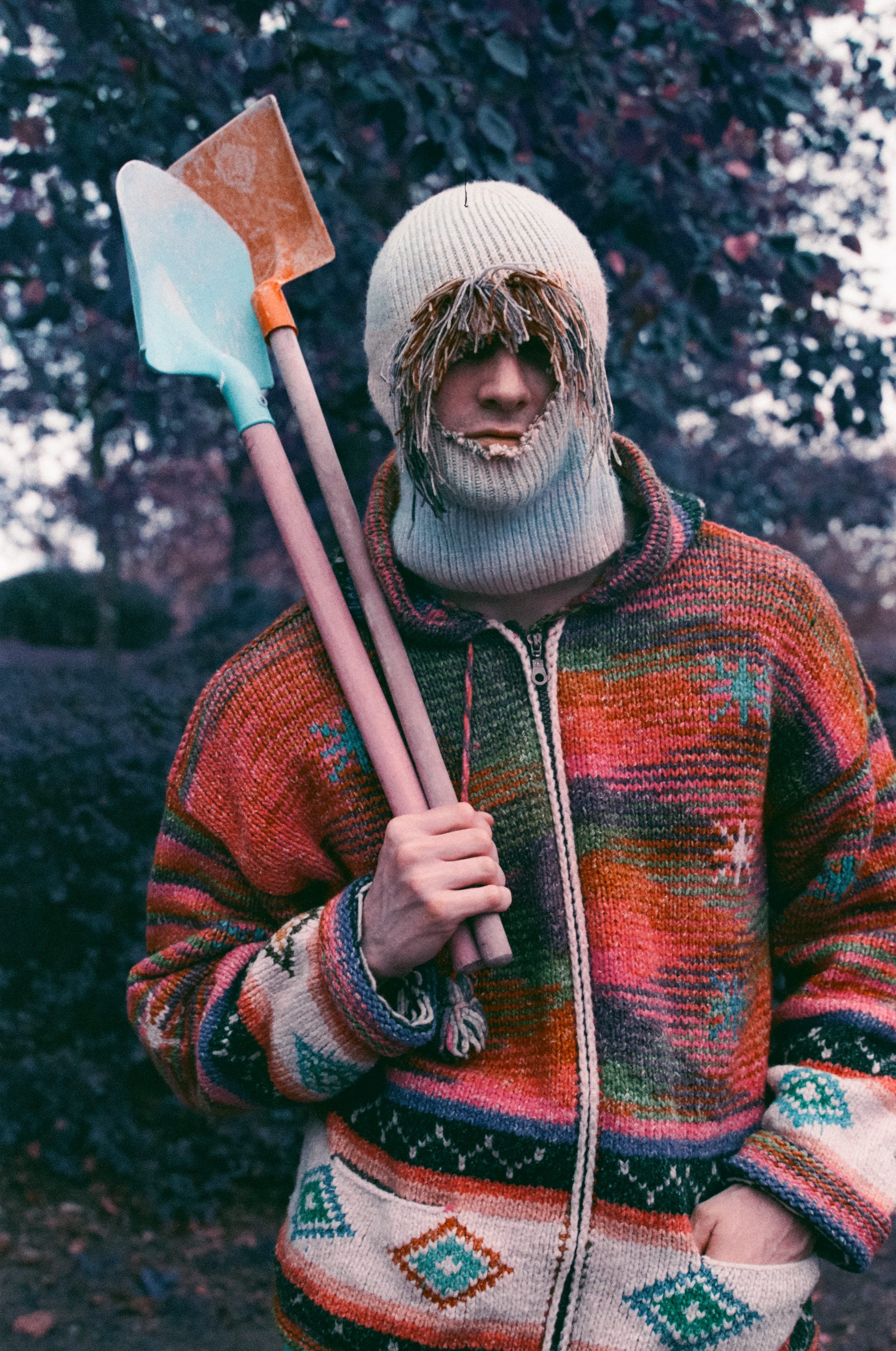 Deux pelles sur l'épaule, une turquoise et une orange. Cagoule écrue frangée, gilet zippé à motifs géométriques, feuillage violet derrière.