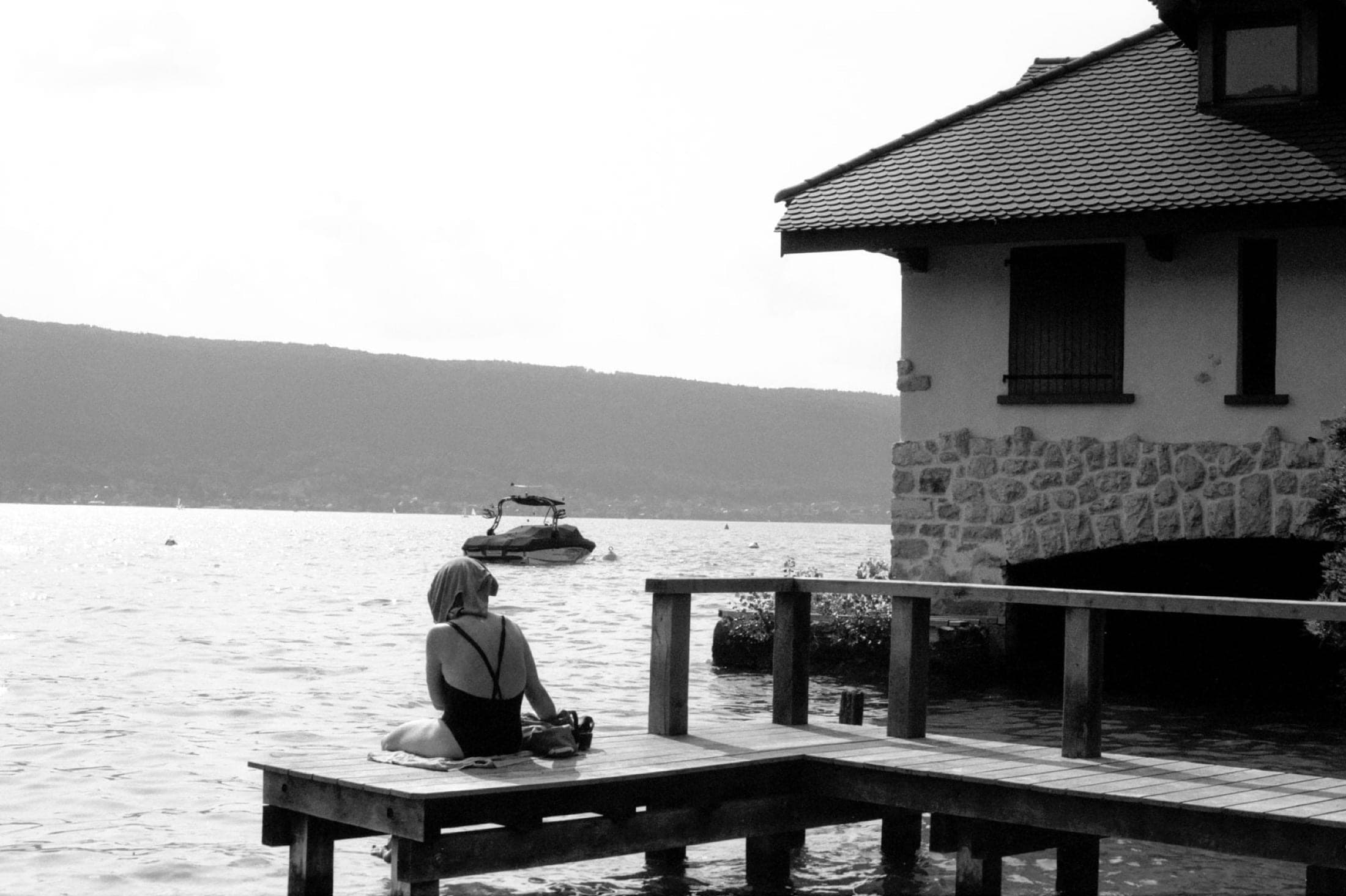 Sur un ponton, une personne en maillot, serviette sur la tête, regarde le lac près d'un hangar à bateaux.