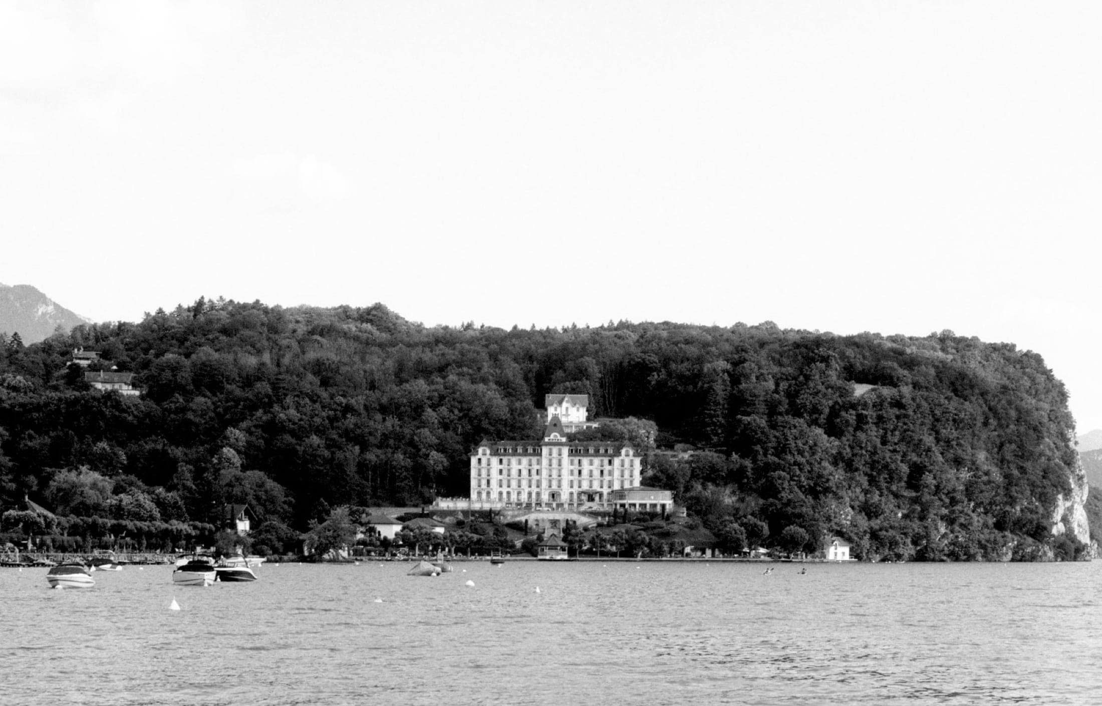 Palace de bord de lac vu depuis l'eau, adossé à une colline boisée, quelques bateaux au mouillage.