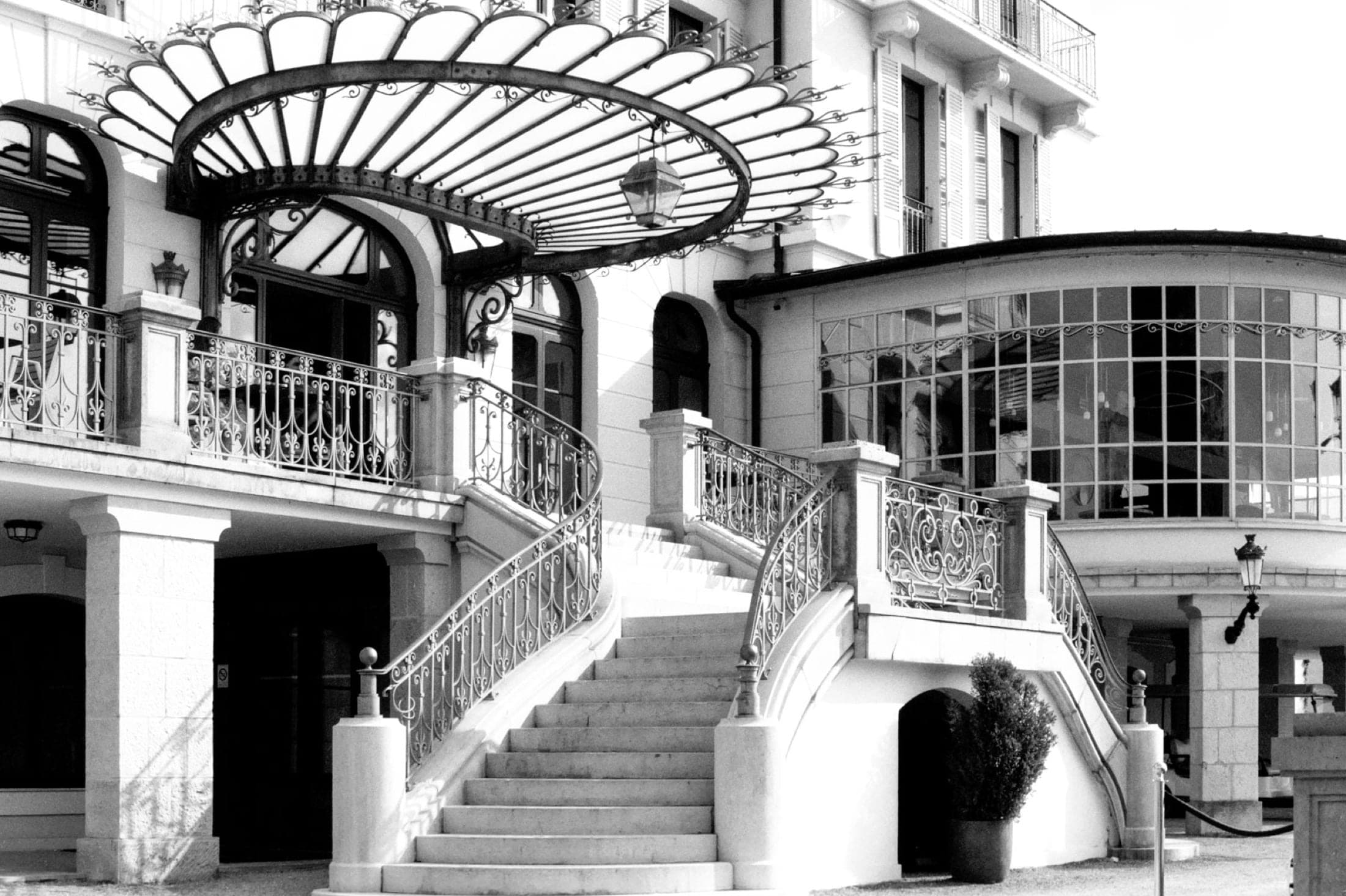 Façade d'un palace Belle Époque : marquise de fer forgé, double escalier de pierre et véranda vitrée.
