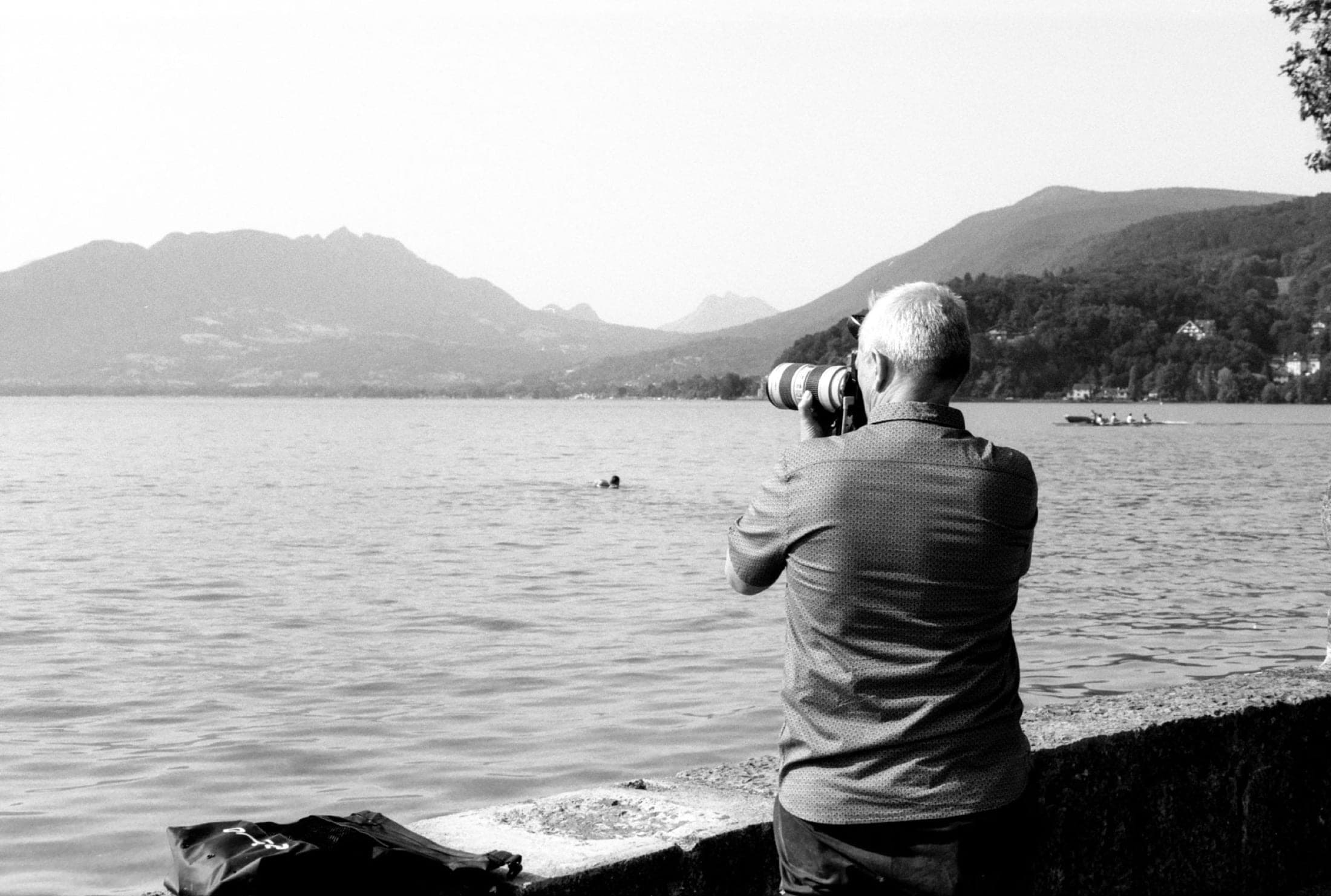 Le photographe, téléobjectif à l'œil, cadre la surface du lac depuis le quai, un aviron au loin.