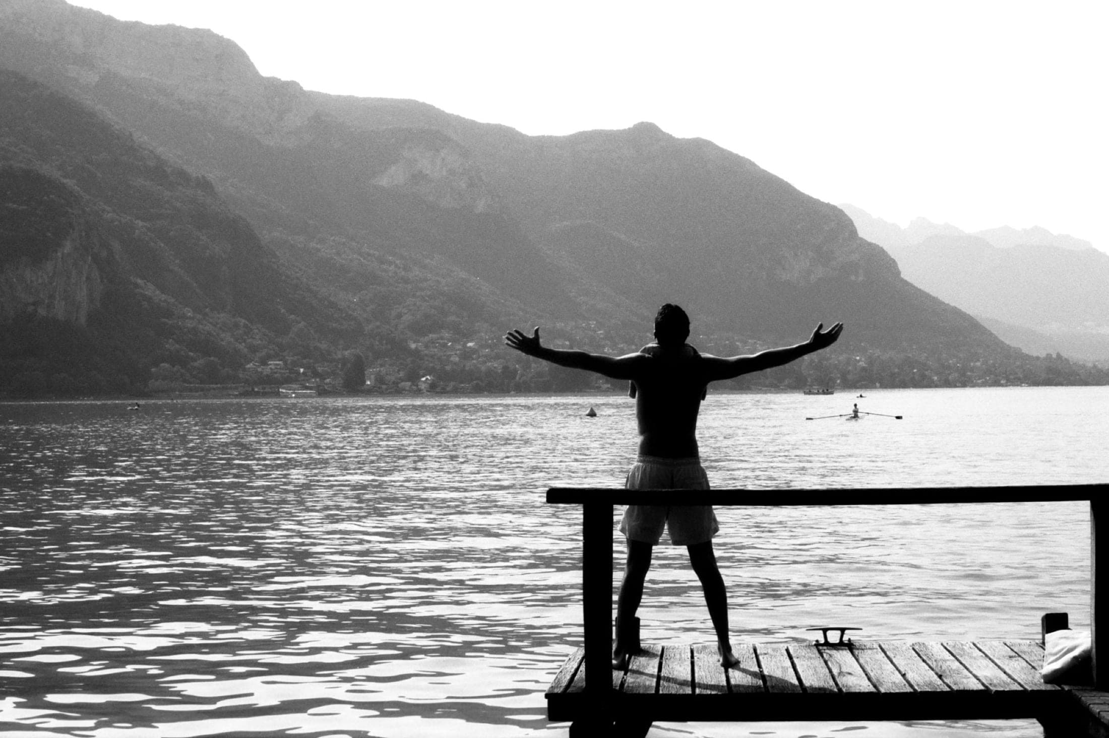 Silhouette à contre-jour sur un ponton, bras grands ouverts face au lac et aux montagnes.