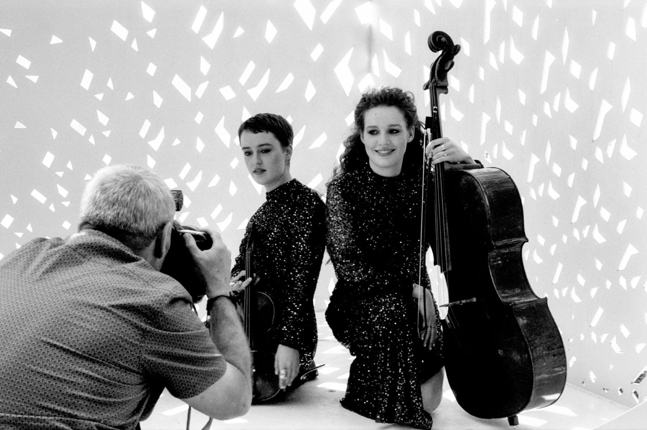 Simon Fowler photographie les sœurs Berthollet agenouillées en robes pailletées, violon et violoncelle en main, devant un fond blanc ajouré.