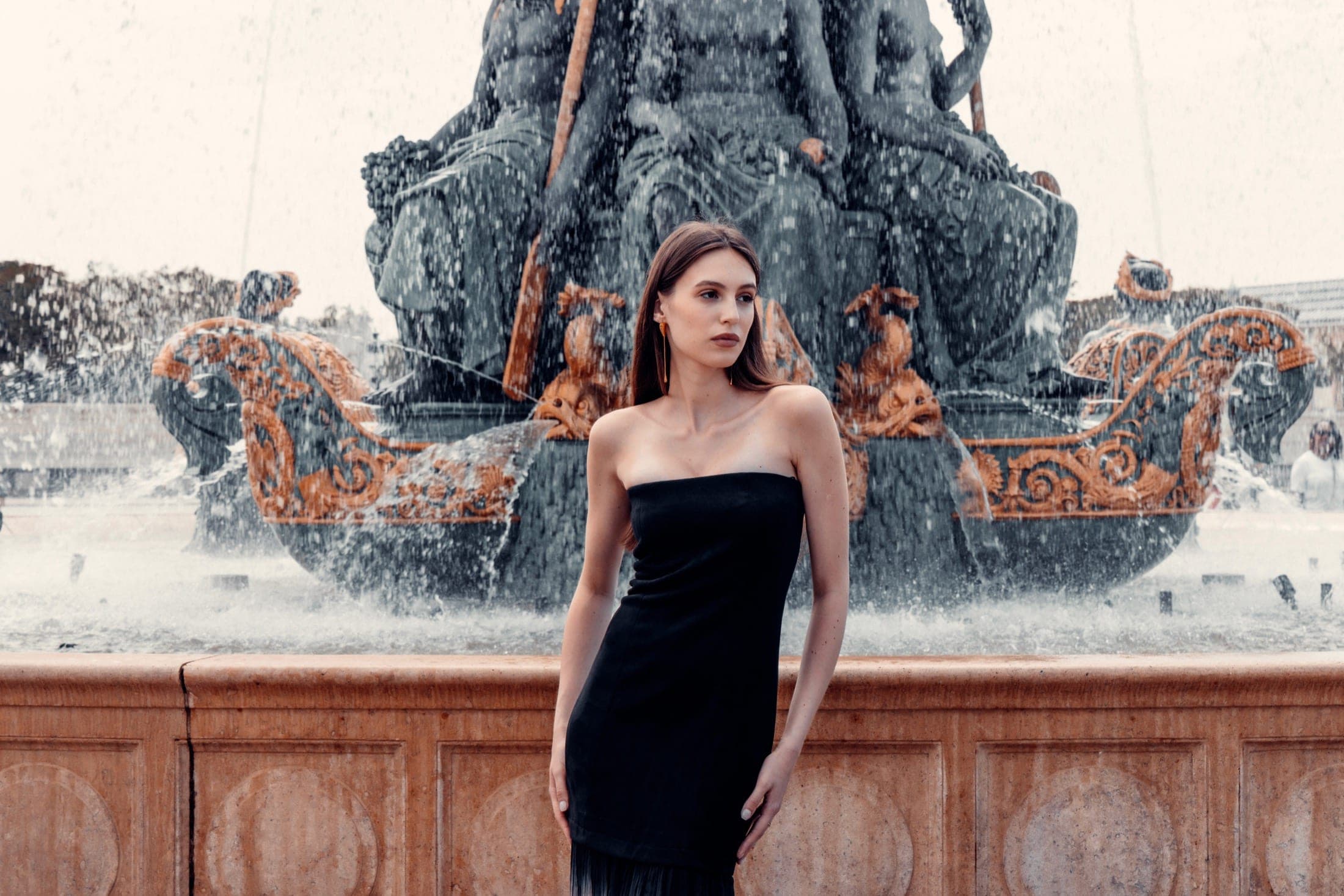 Grand angle sur la robe noire devant la fontaine, le regard tourné de côté, statue et jets d'eau pleinement visibles.