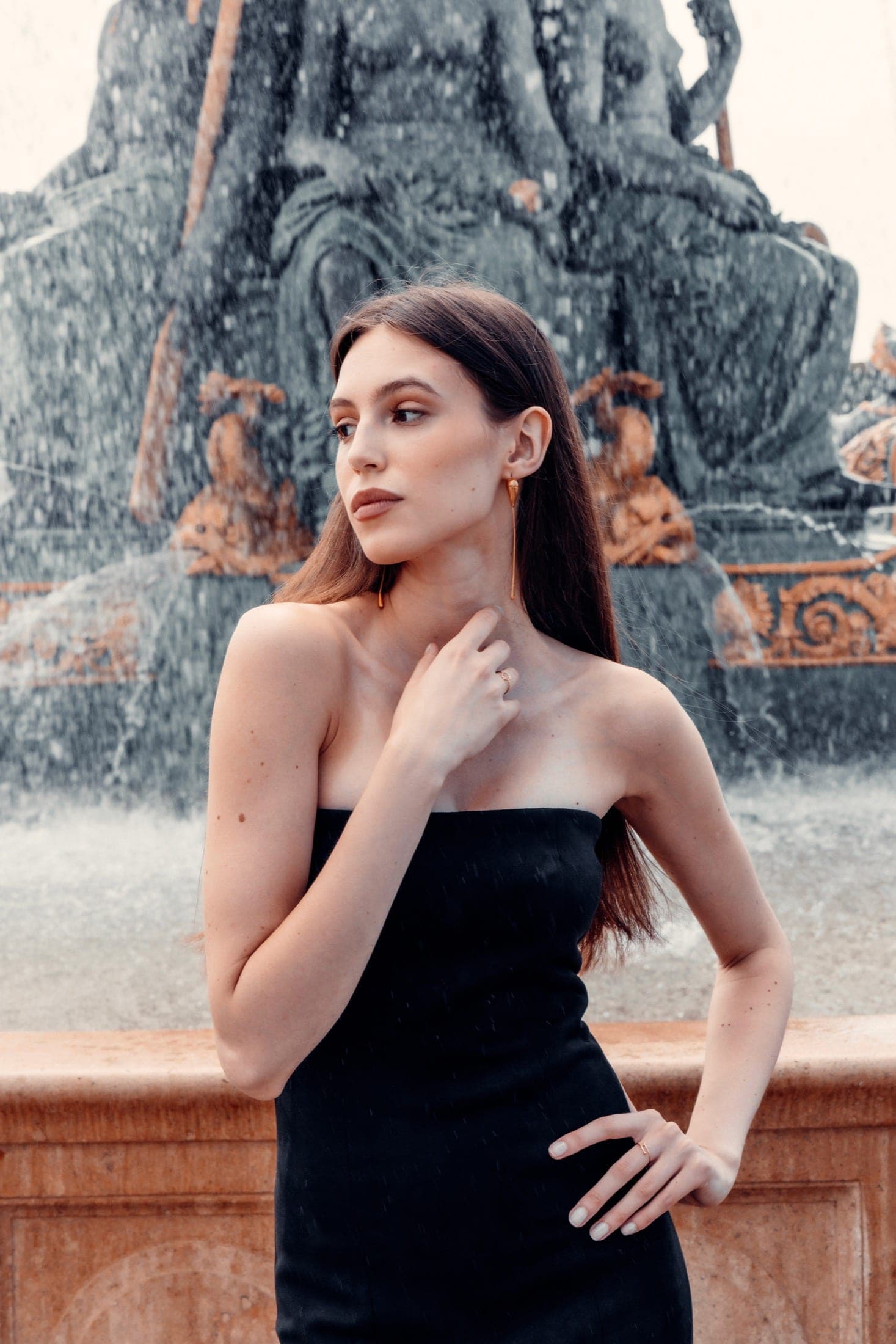 Une main posée sur la nuque, boucles d'oreilles pendantes, la fontaine floutée derrière.