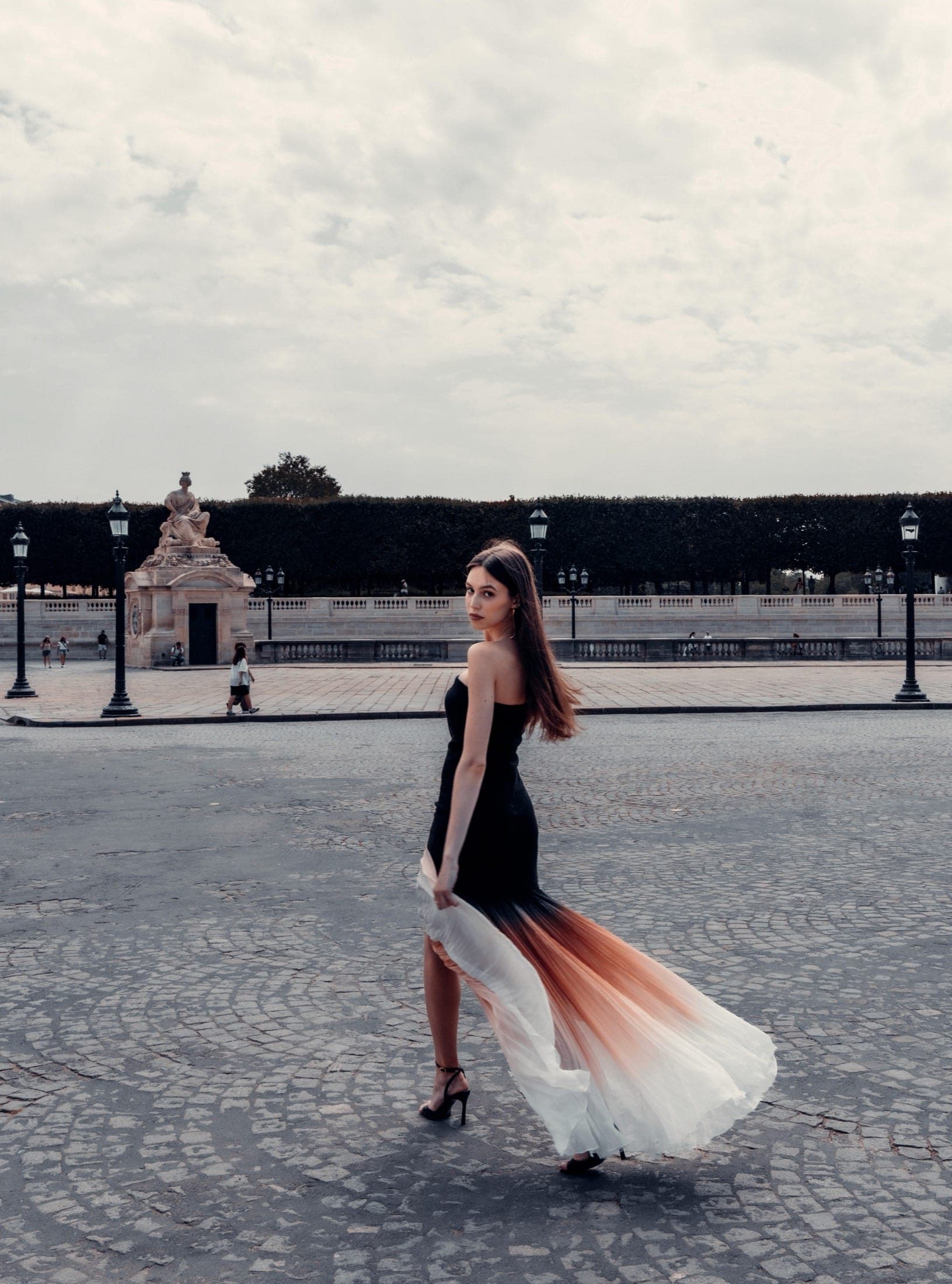 La jupe plissée dégradée en mouvement à travers la place de la Concorde, le regard tourné vers l'objectif, statue et lampadaires au loin.