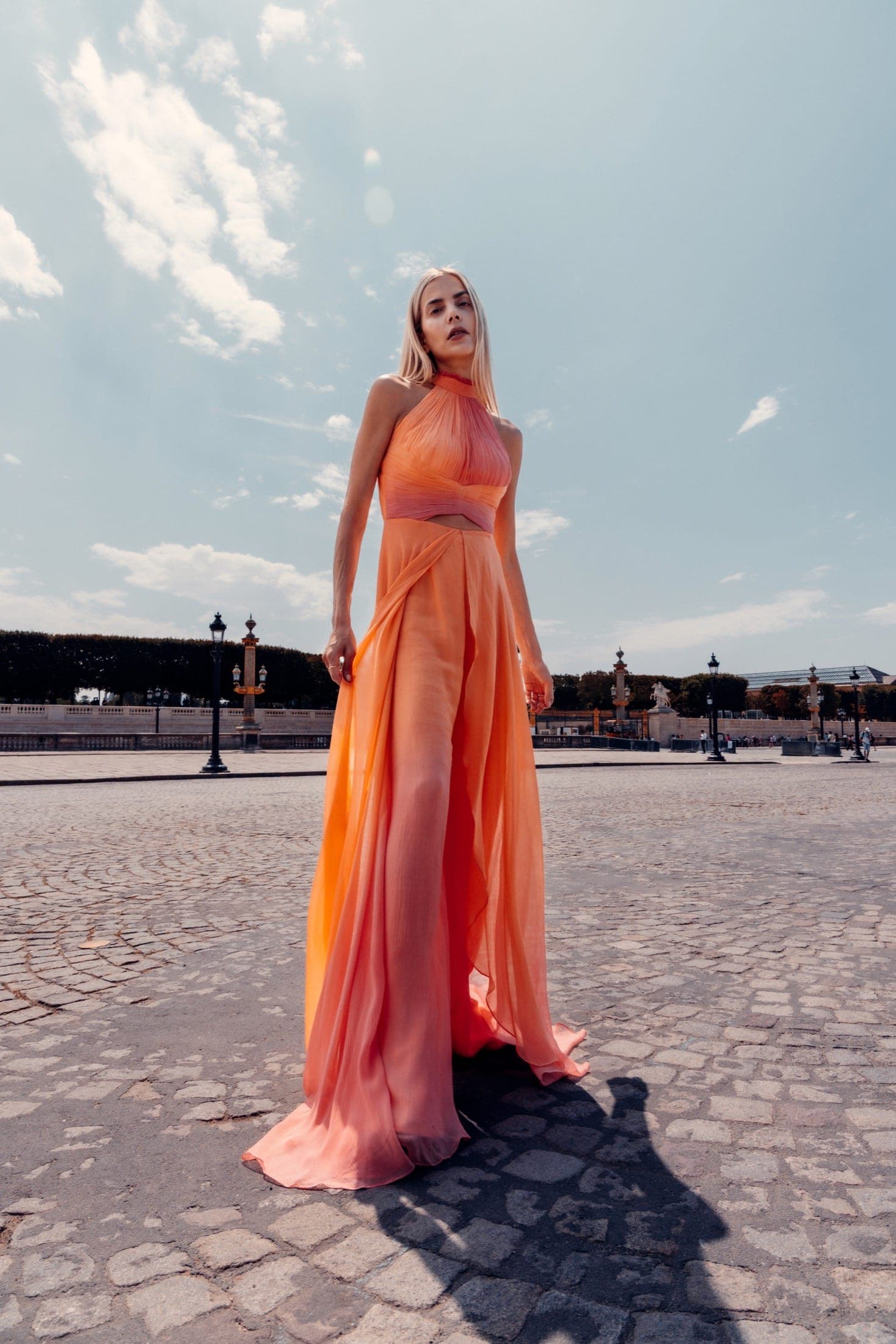 Robe longue dos-nu orange corail à col halter, drapée, debout sur les pavés en plein soleil.