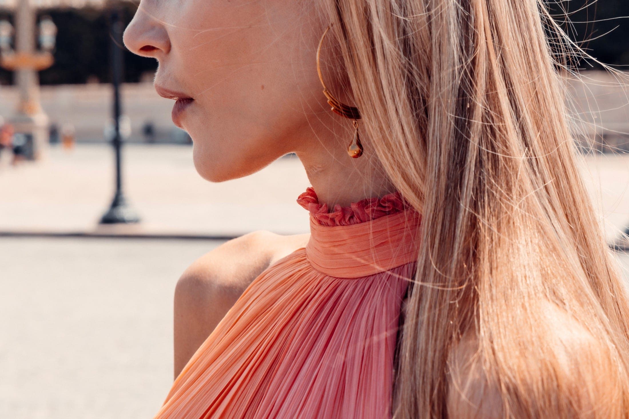 Col plissé orange-rose de la robe halter en gros plan, boucle d'oreille pendante, mèches soulevées par le vent.
