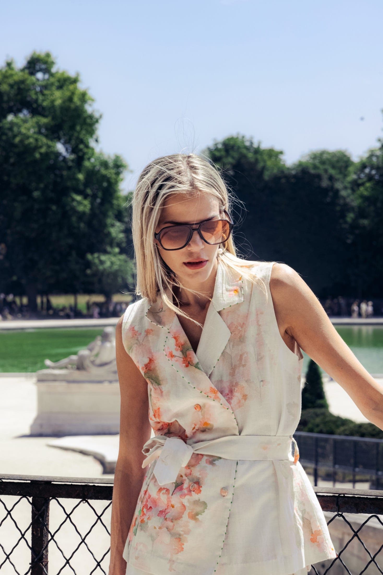 Buste du même gilet fleuri en plan rapproché, lunettes aviateur, un bras tendu appuyé sur la balustrade.