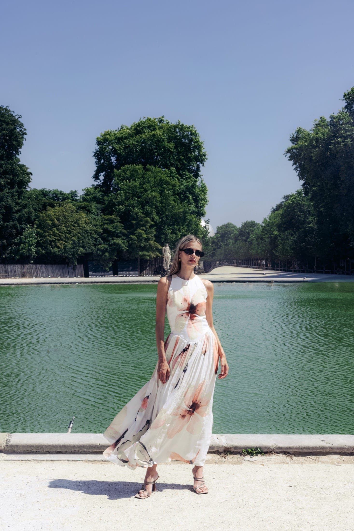 Robe blanche à motif floral et lunettes fines, une main sur la hanche, bassin et allée d'arbres des Tuileries en arrière-plan.