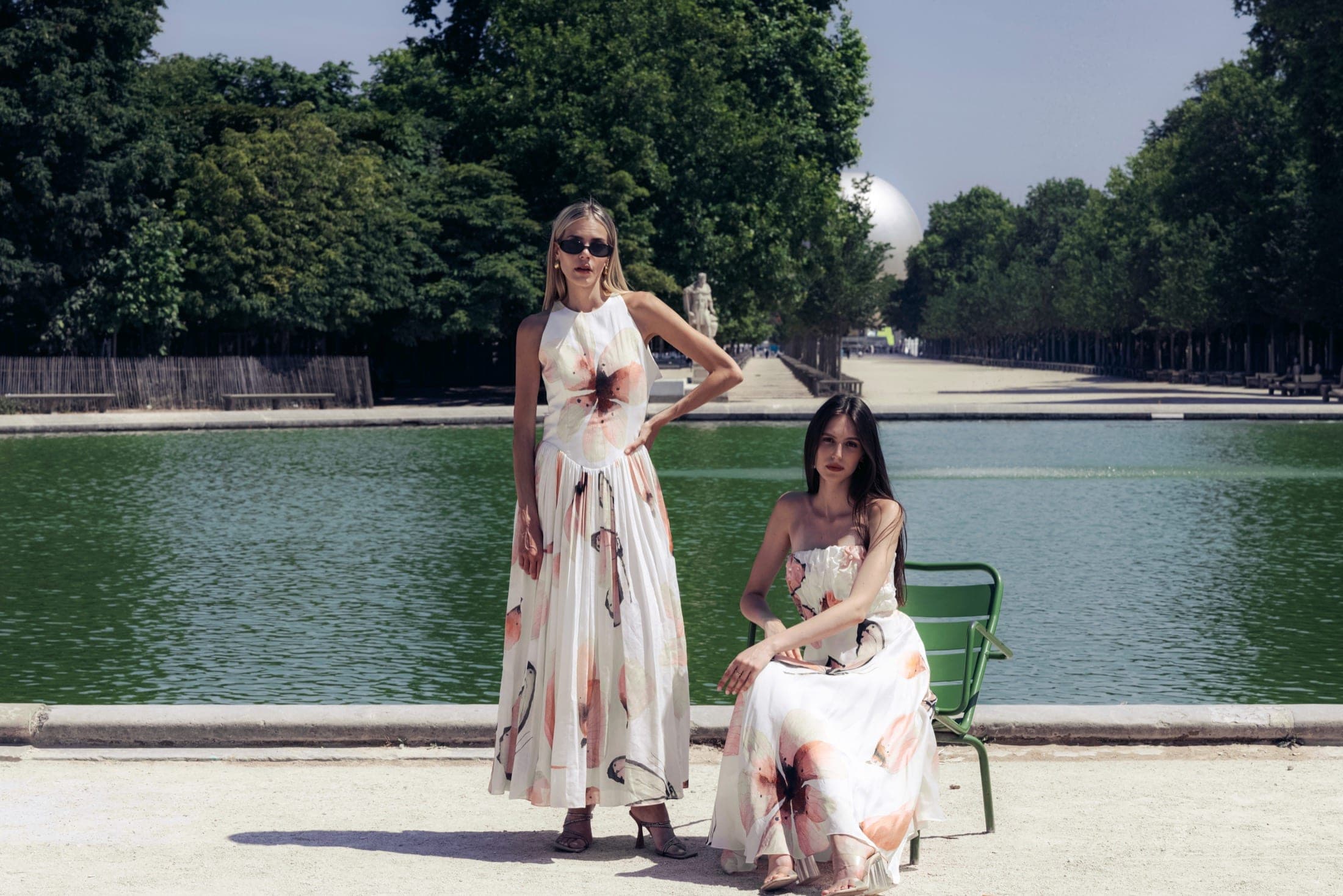 Grand angle sur les deux mannequins devant le bassin, robes longues à motif floral, l'une assise jambes croisées.