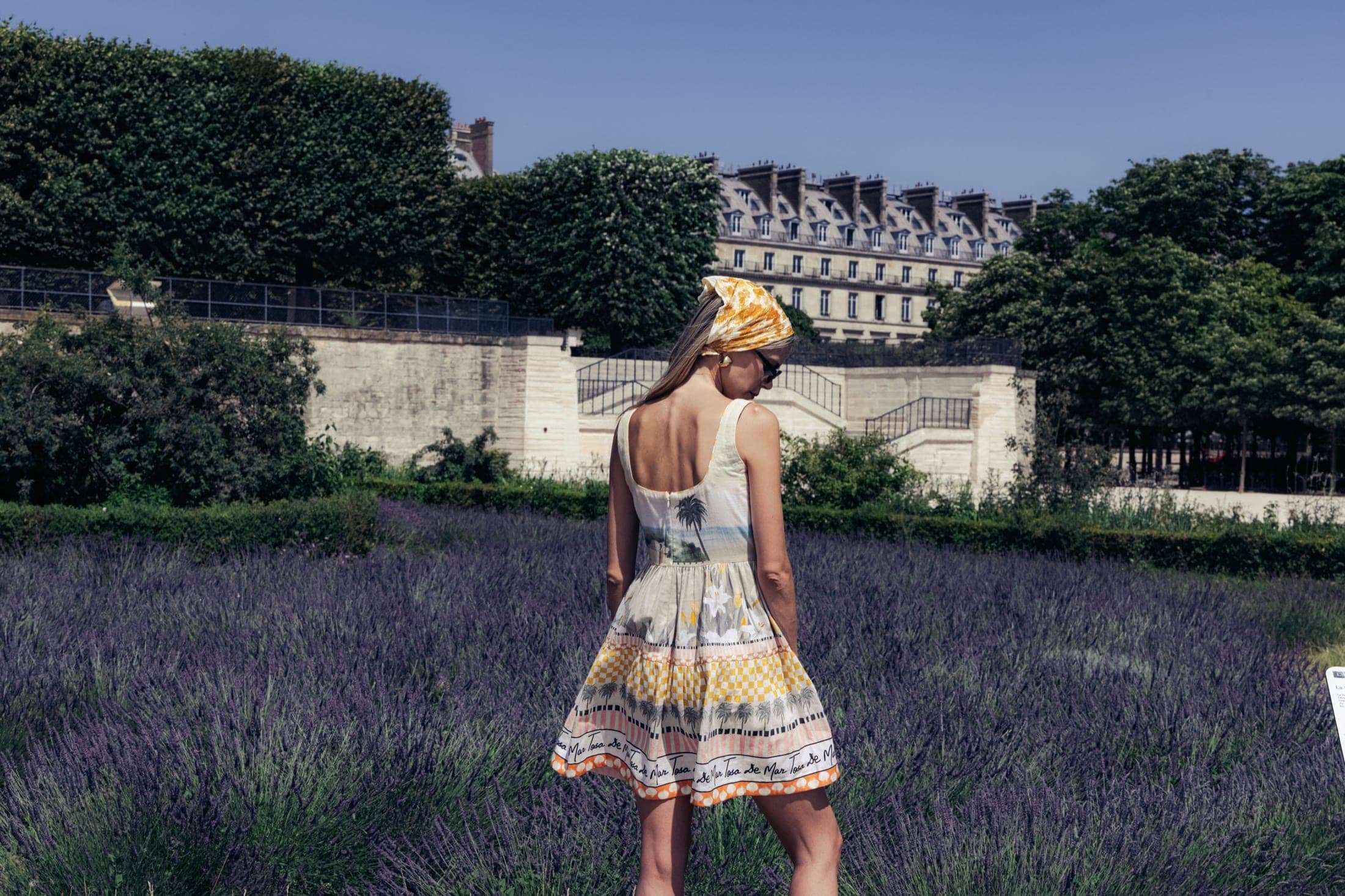 De dos, foulard noué en bandana jaune marbré, robe à motif palmiers et scène balnéaire, champ de lavande et façade parisienne en arrière-plan.