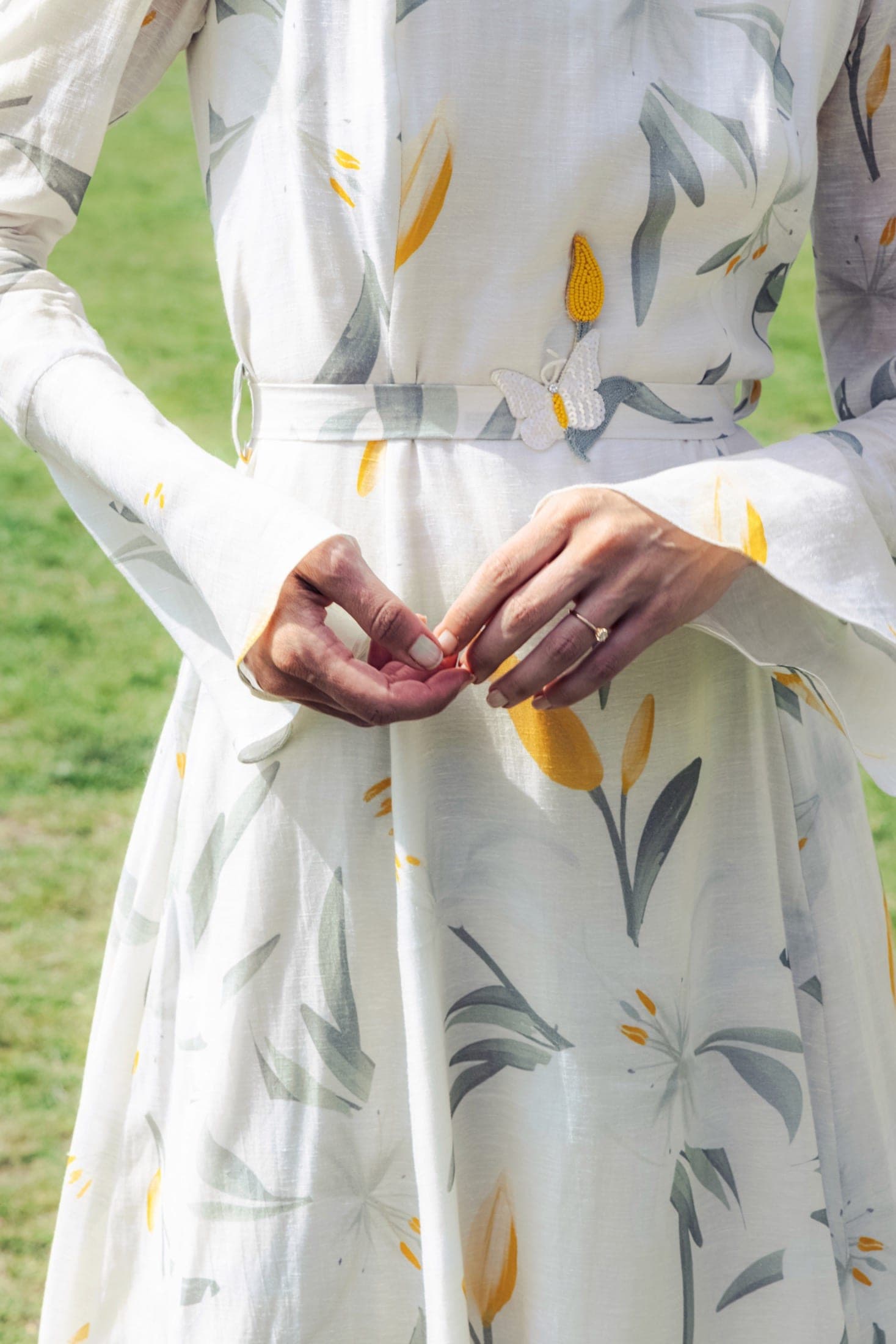 De dos, robe blanche à motif de tulipes jaune et gris, broche papillon perlée à la taille, mains jointes dans le dos.