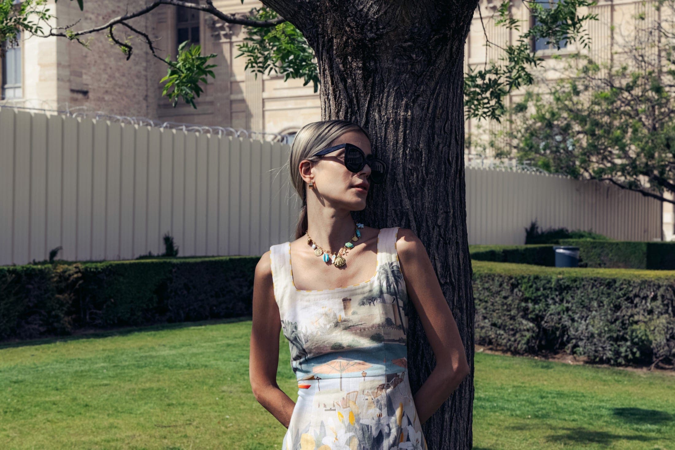 Debout adossée au même tronc, de profil, lunettes de soleil et collier de coquillages, ensemble à motif balnéaire.