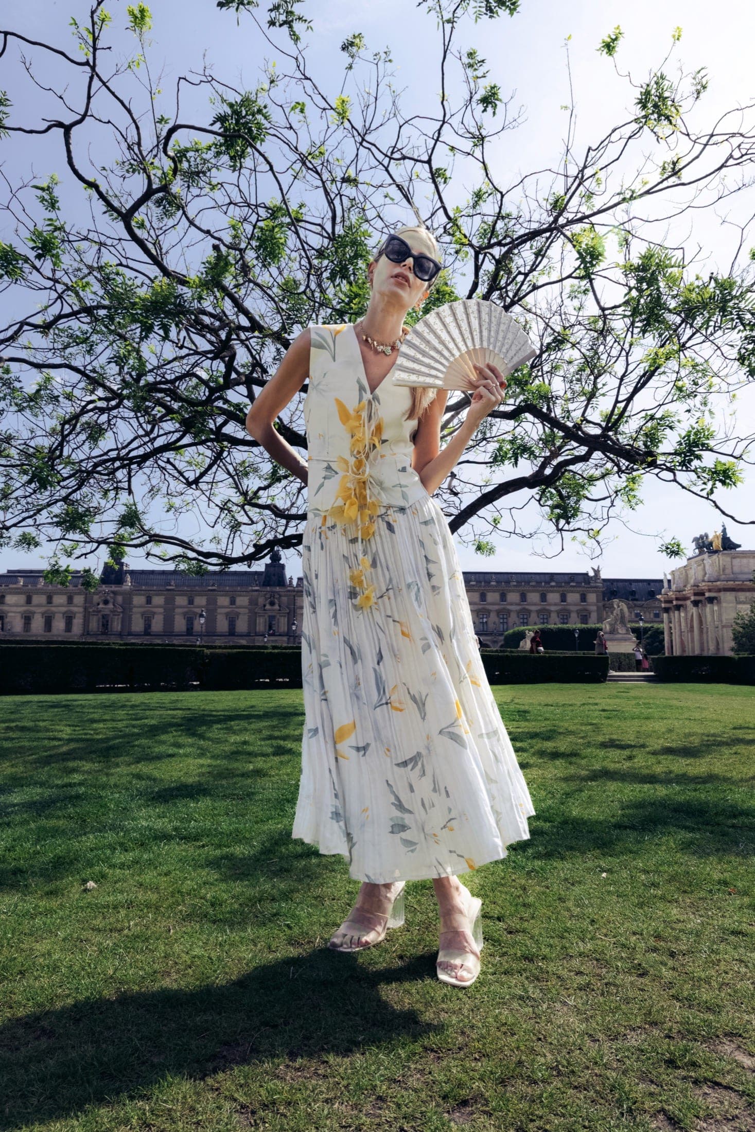 Robe longue plissée à motif de tulipes jaune et gris, éventail en tissu brodé blanc à la main, cour du Carrousel du Louvre en arrière-plan.