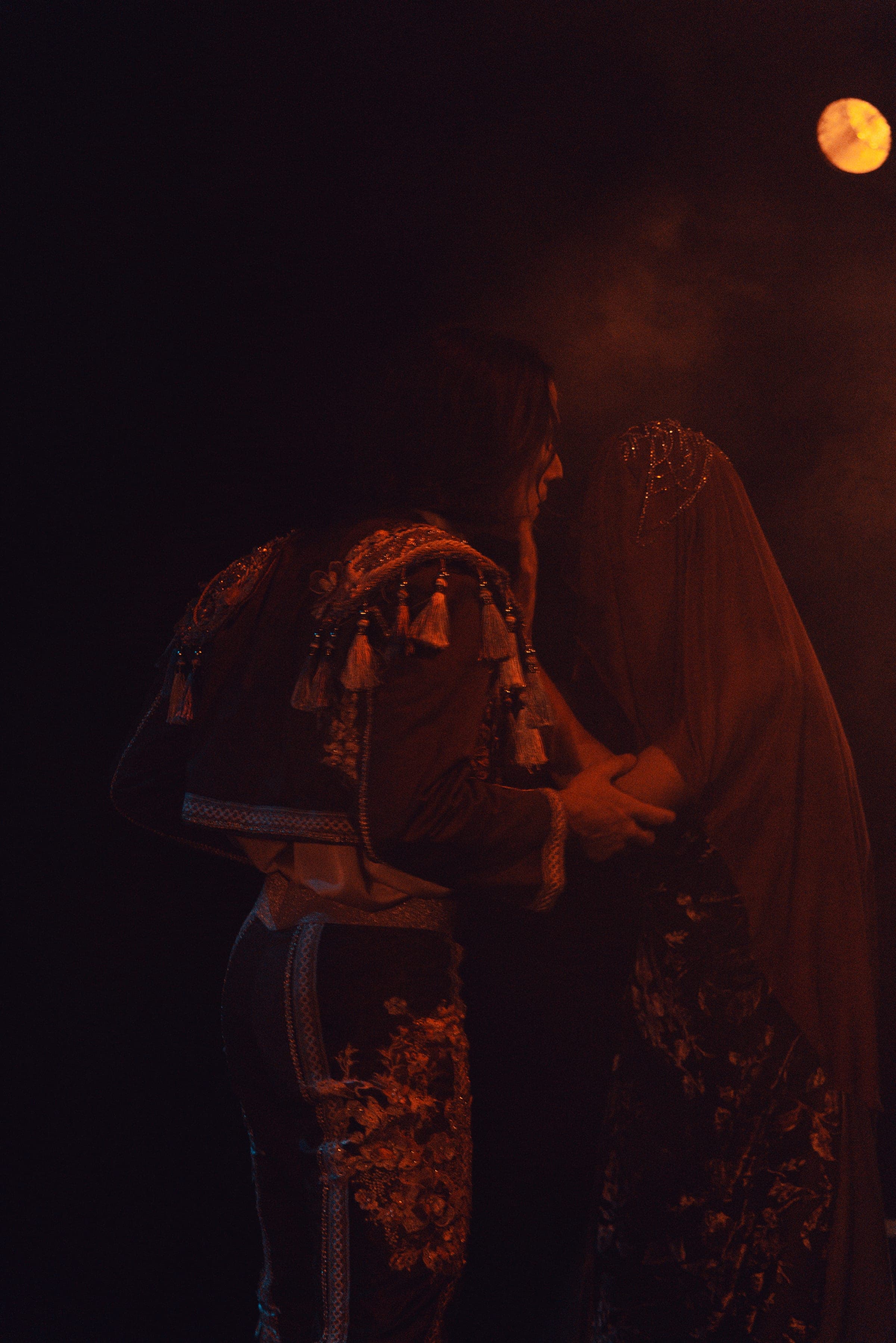 Deux personnages de dos sous une lune de théâtre, voile brodé et lumière rouge