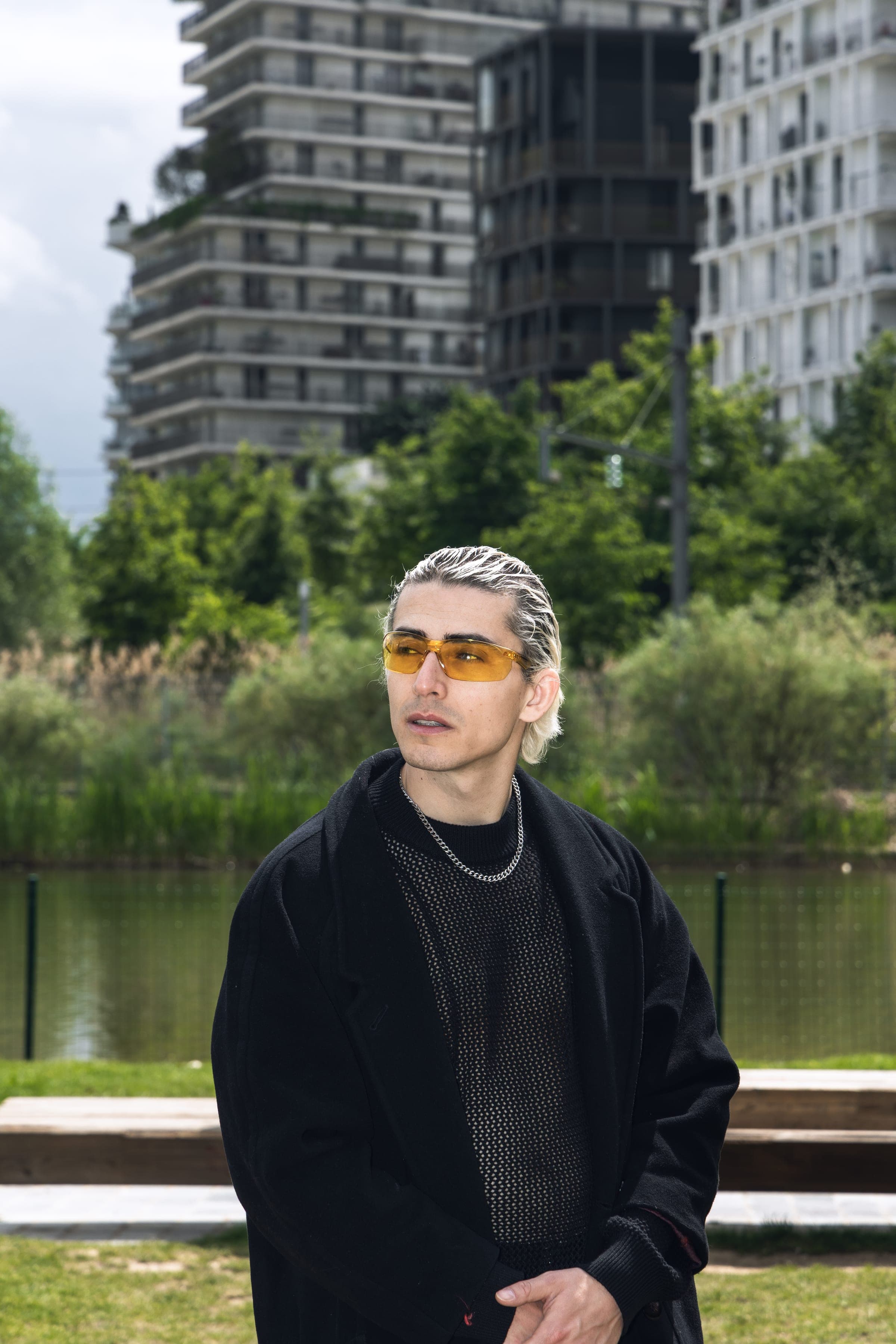 Portrait devant un plan d'eau, lunettes jaunes et chaîne argentée, les tours au fond.