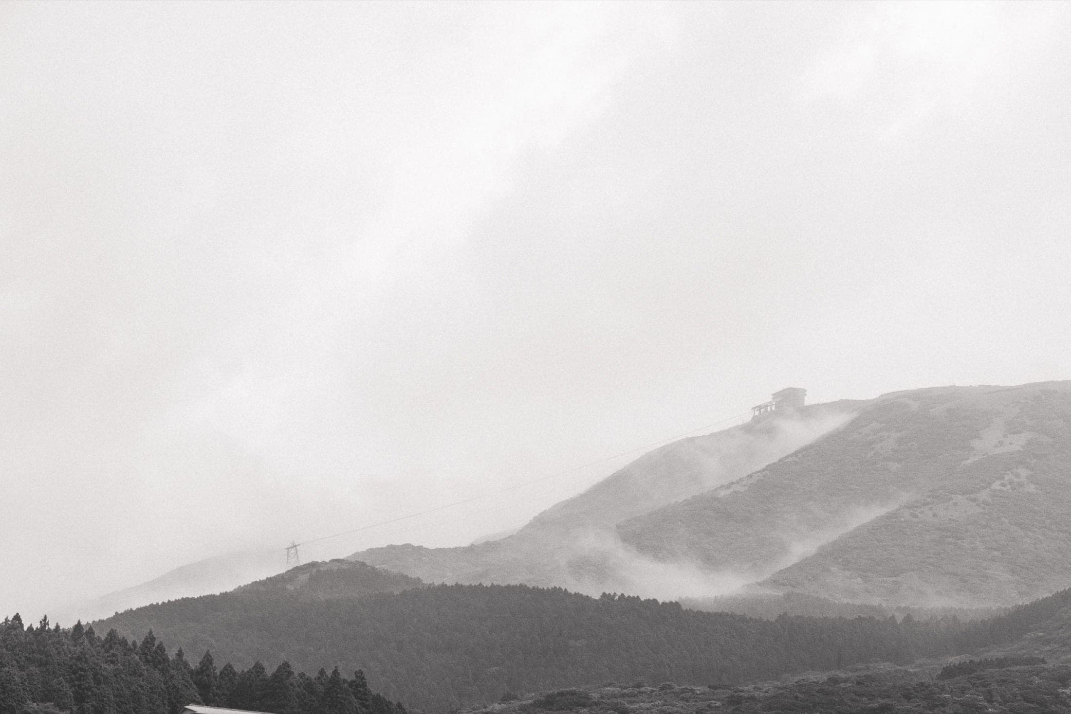 Noir et blanc : crêtes brumeuses, une station de téléphérique posée sur la ligne de crête, câbles tendus au-dessus des forêts.