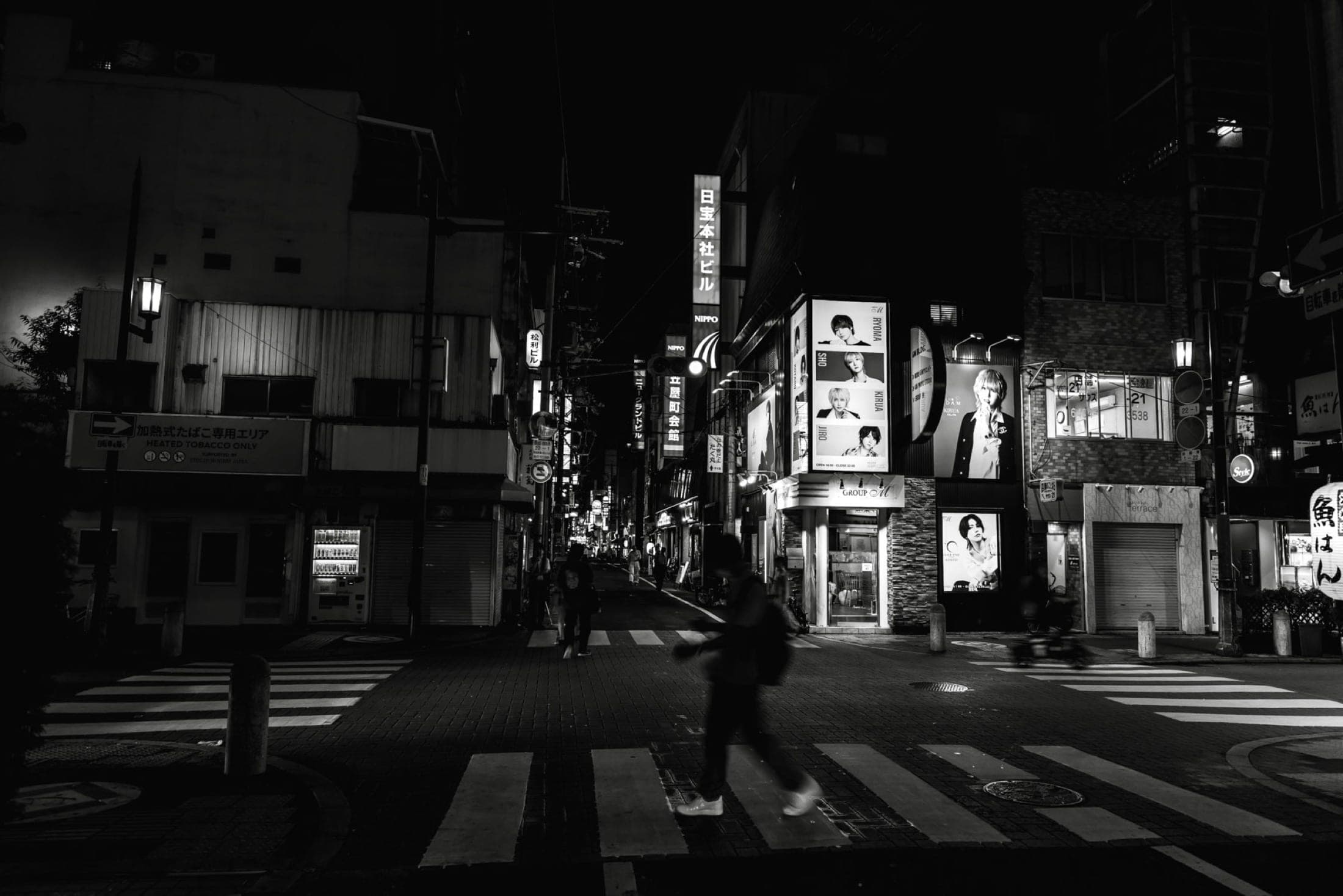 Noir et blanc : carrefour désert la nuit, panneaux publicitaires de clubs, distributeur automatique, un passant flou sur le passage piéton.