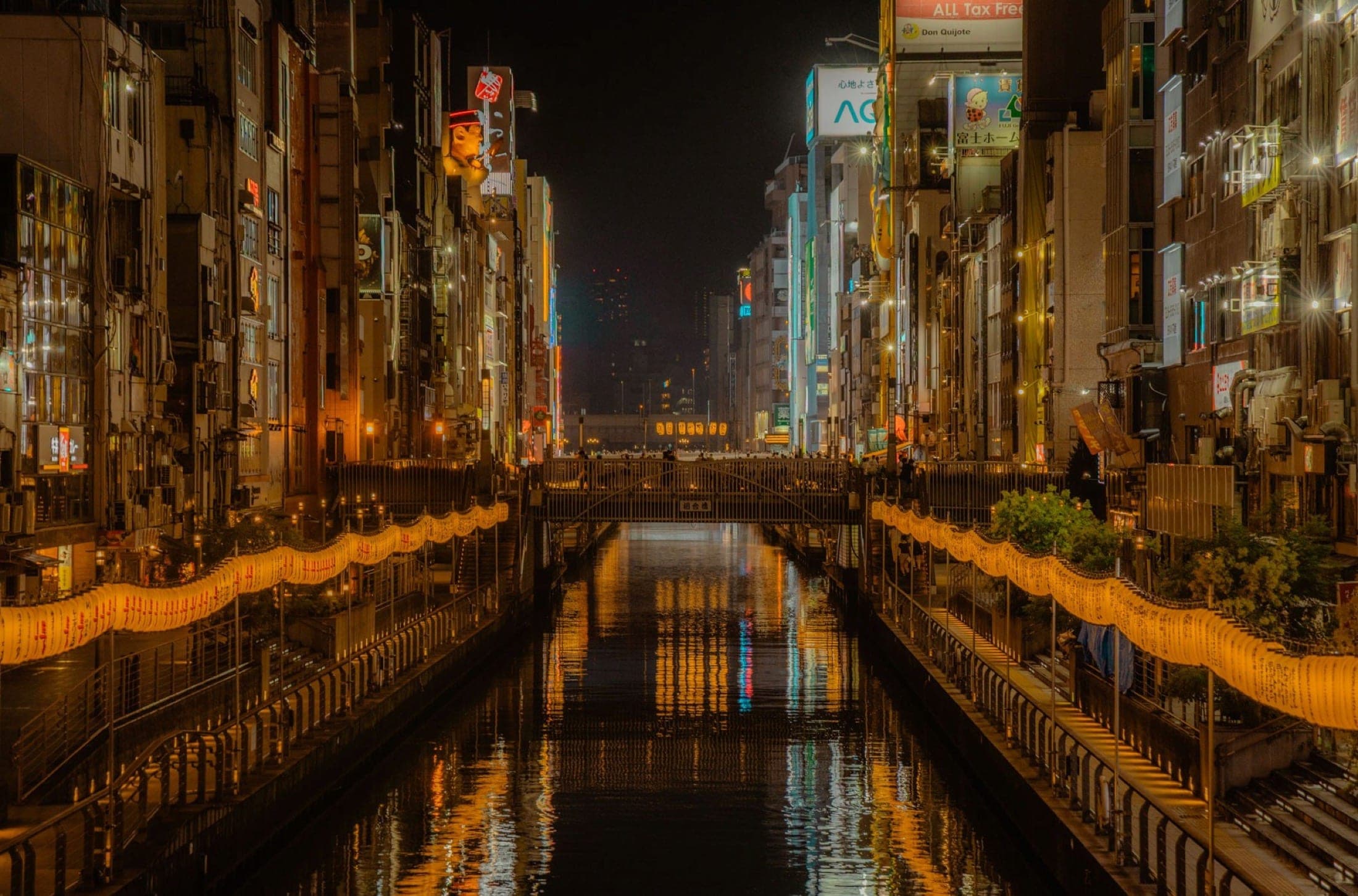 Canal urbain la nuit, guirlandes de lanternes le long des deux rives et enseignes lumineuses se reflétant dans l'eau noire.
