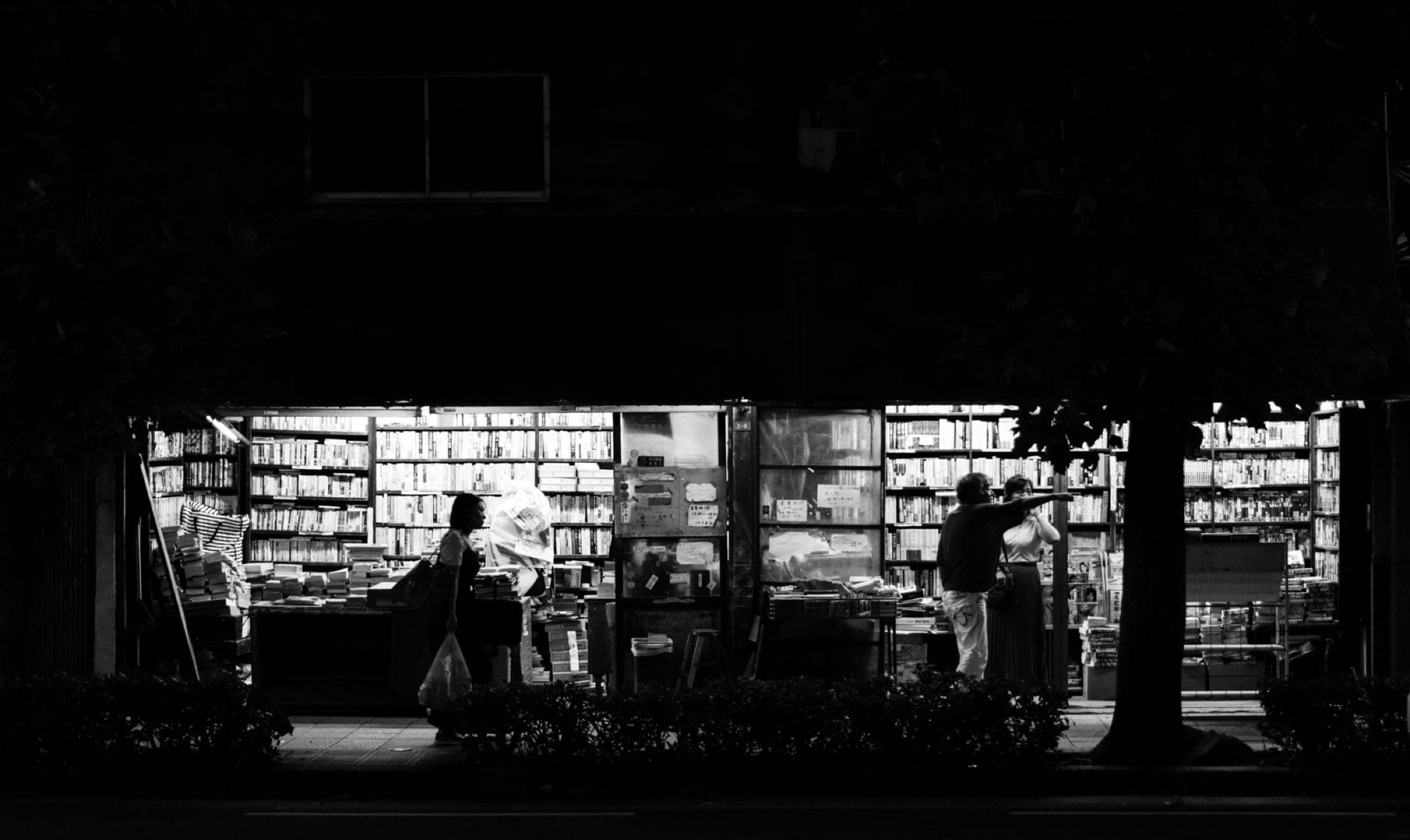 Noir et blanc : librairie d'occasion ouverte sur la rue la nuit, murs de livres éclairés, silhouettes de clients devant les étals.