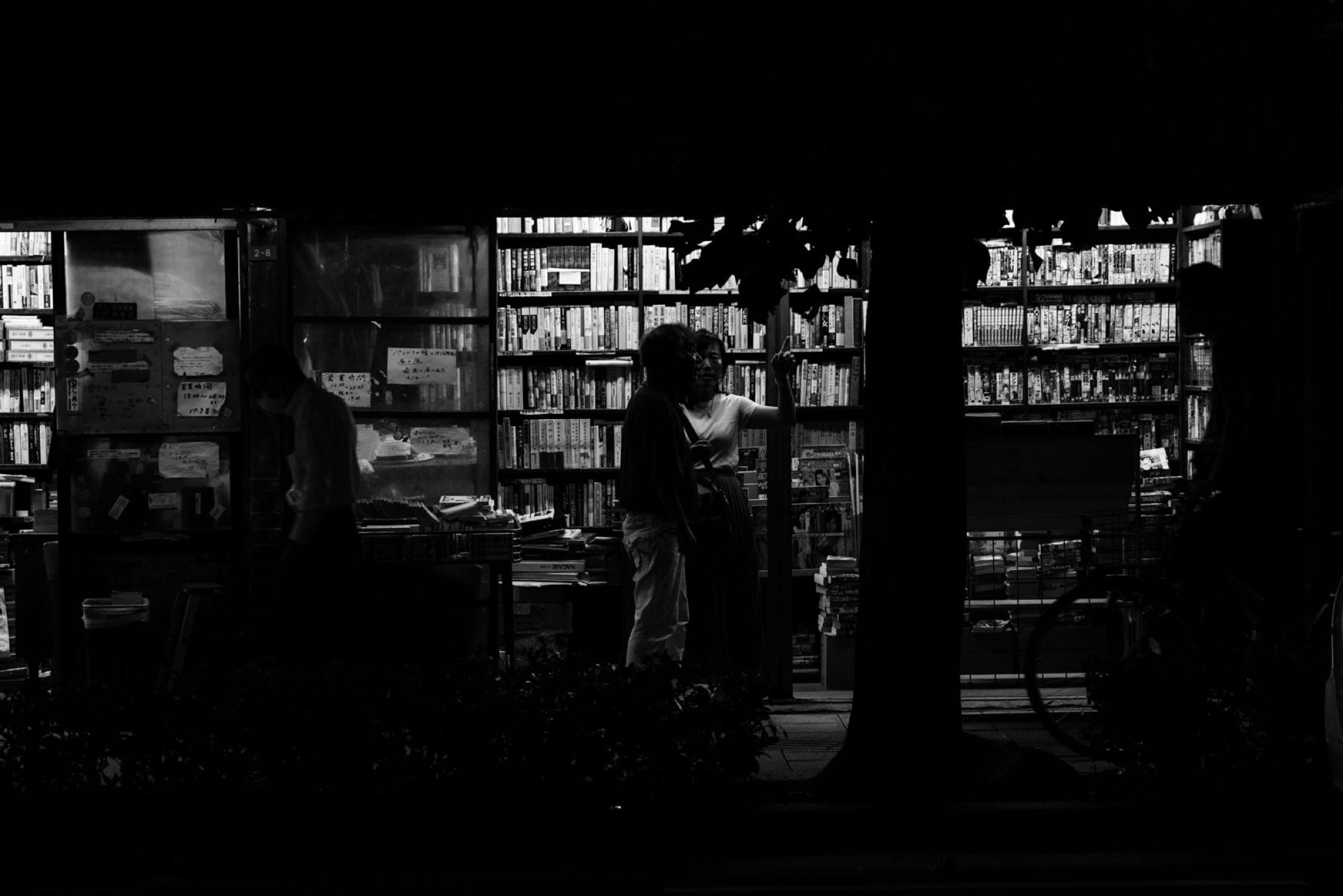 Noir et blanc : devant les rayonnages d'une librairie de nuit, deux personnes désignent un livre pendant qu'une troisième passe derrière la vitre.