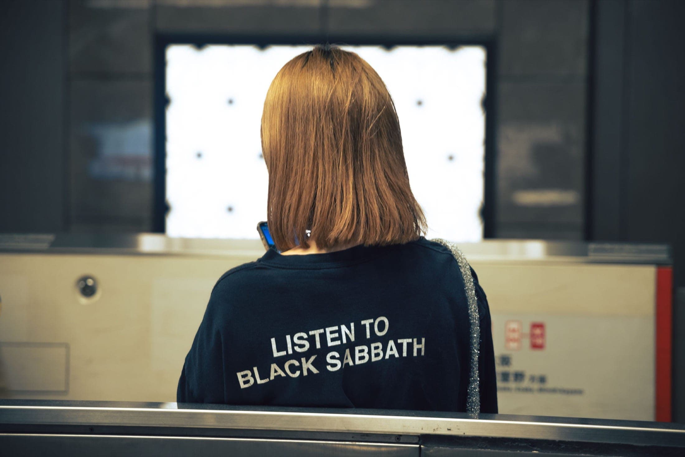 De dos dans une gare, une personne en sweat noir portant l'inscription Listen to Black Sabbath, téléphone à la main.
