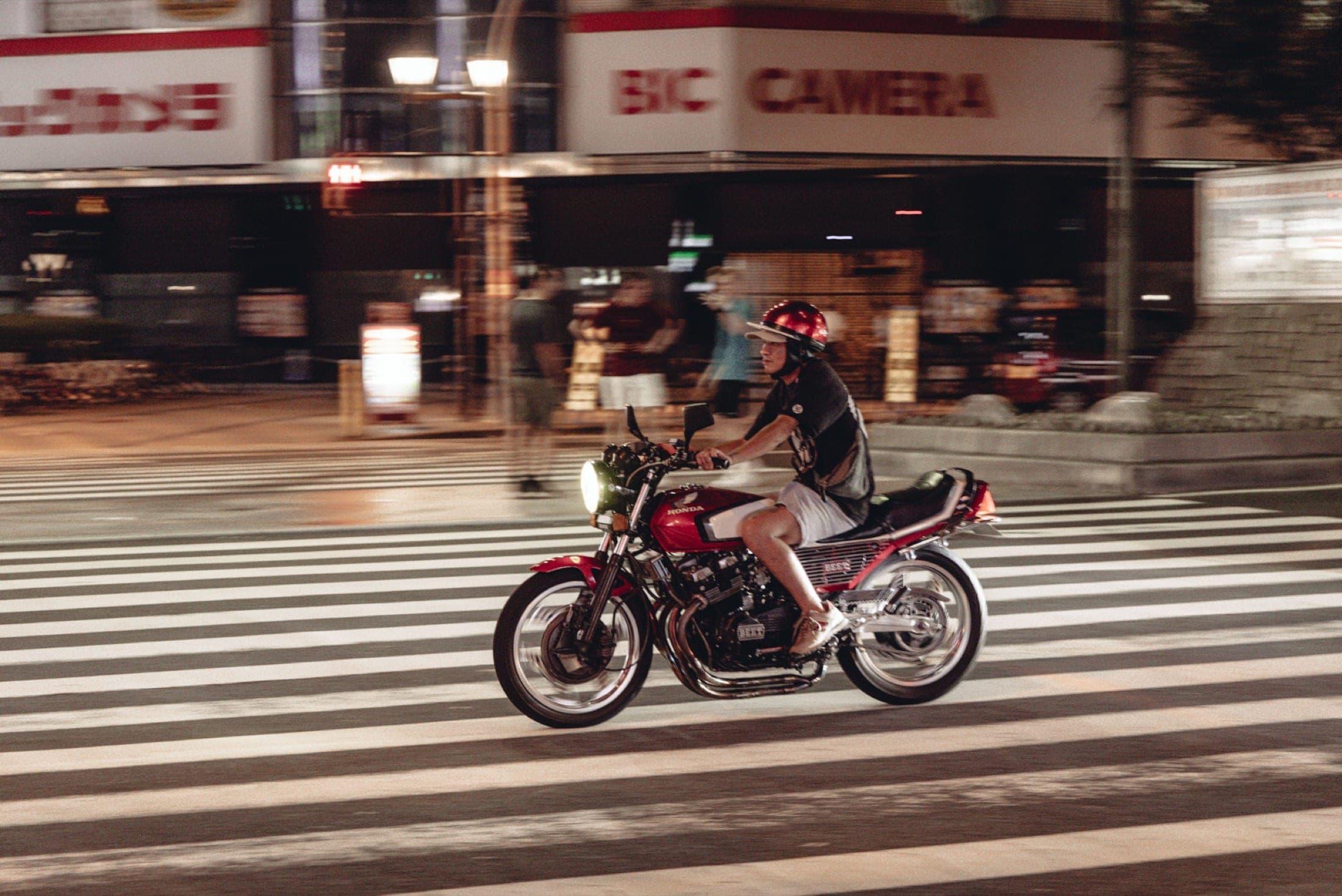 Filé sur un motard casqué de rouge traversant un passage piéton, la façade d'un grand magasin d'électronique floue derrière.