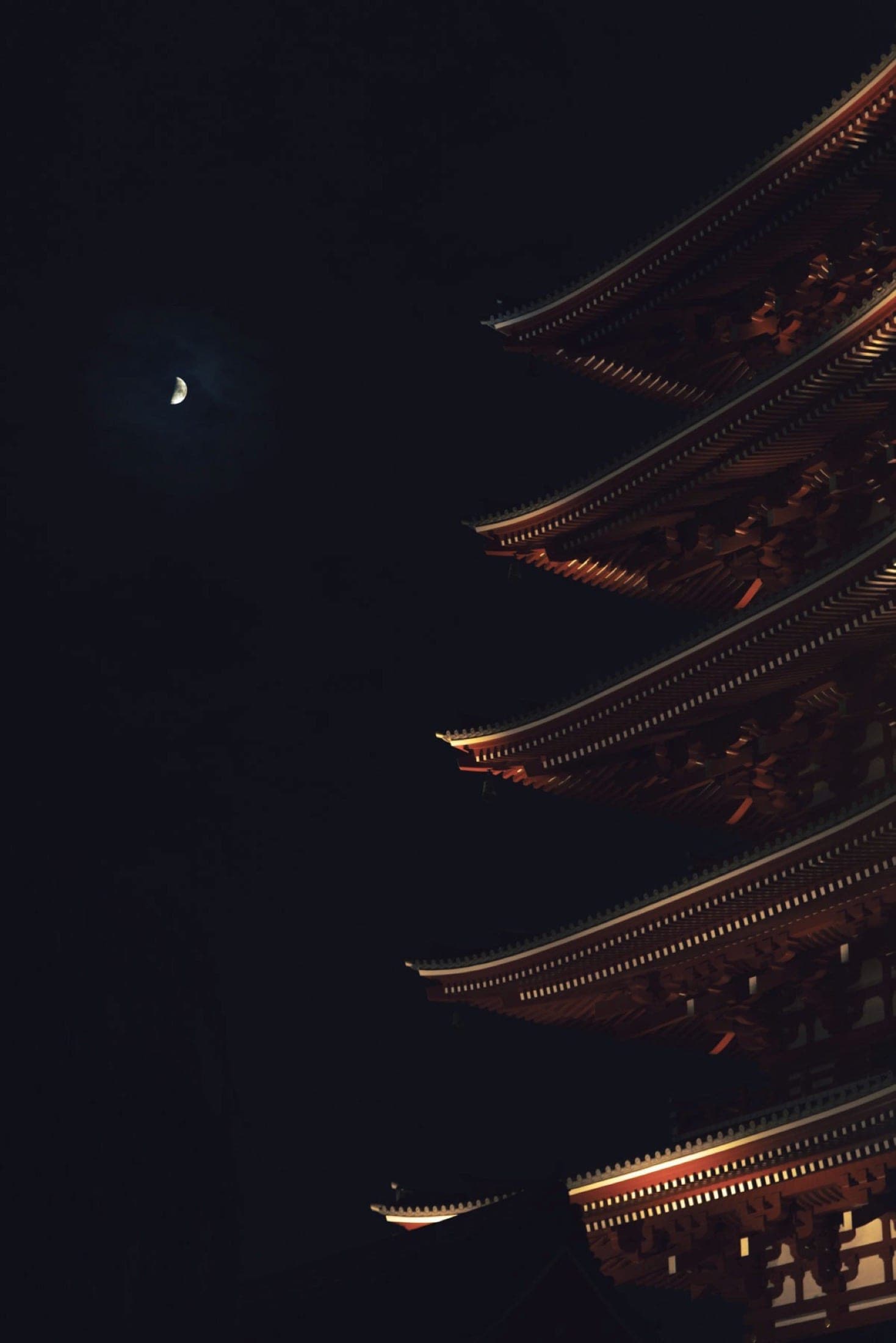 Détail des avant-toits superposés d'une pagode éclairée, un croissant de lune dans le ciel noir à gauche.