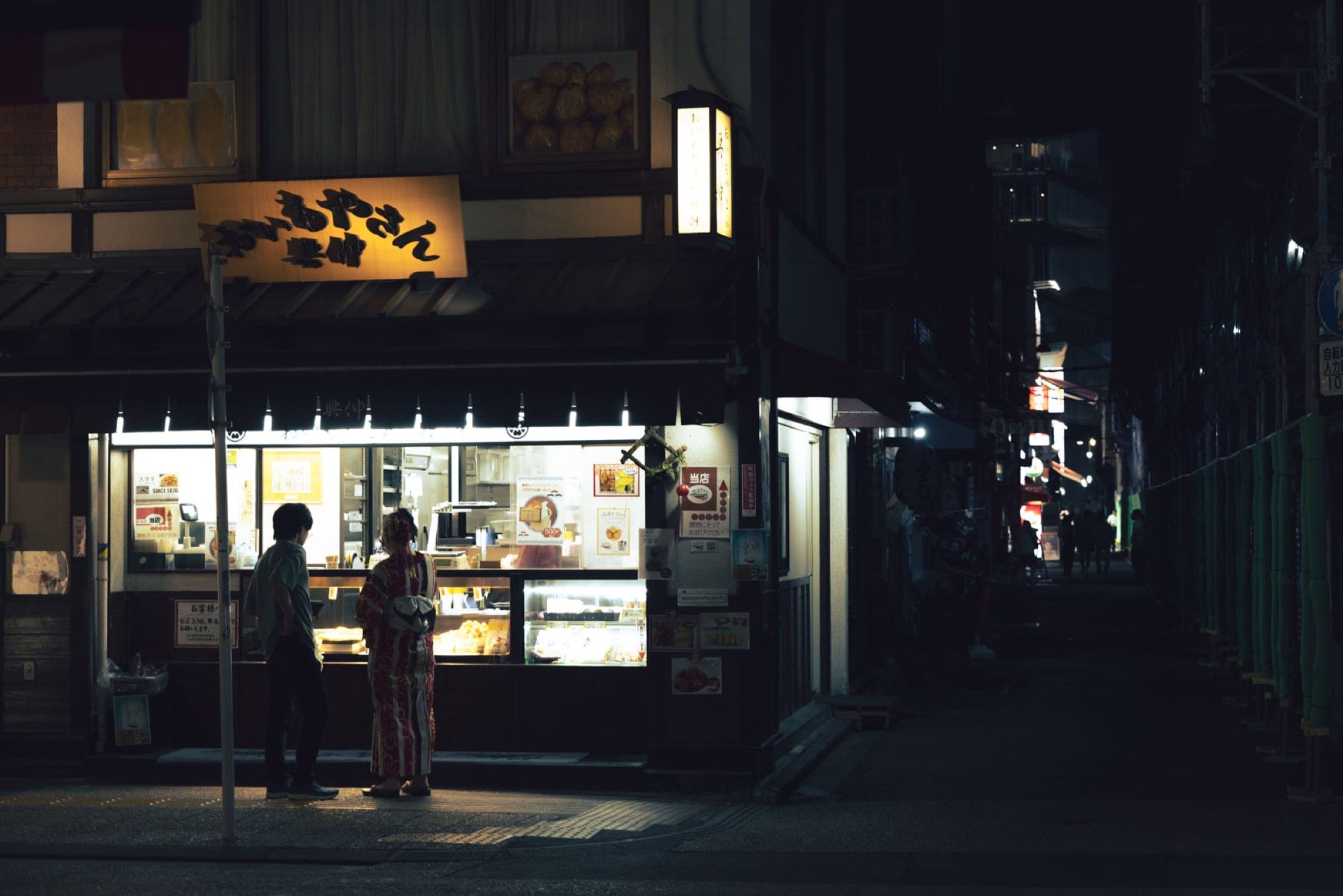 Deux personnes devant la vitrine éclairée d'une confiserie la nuit, l'une en yukata à motifs, ruelle sombre à droite.