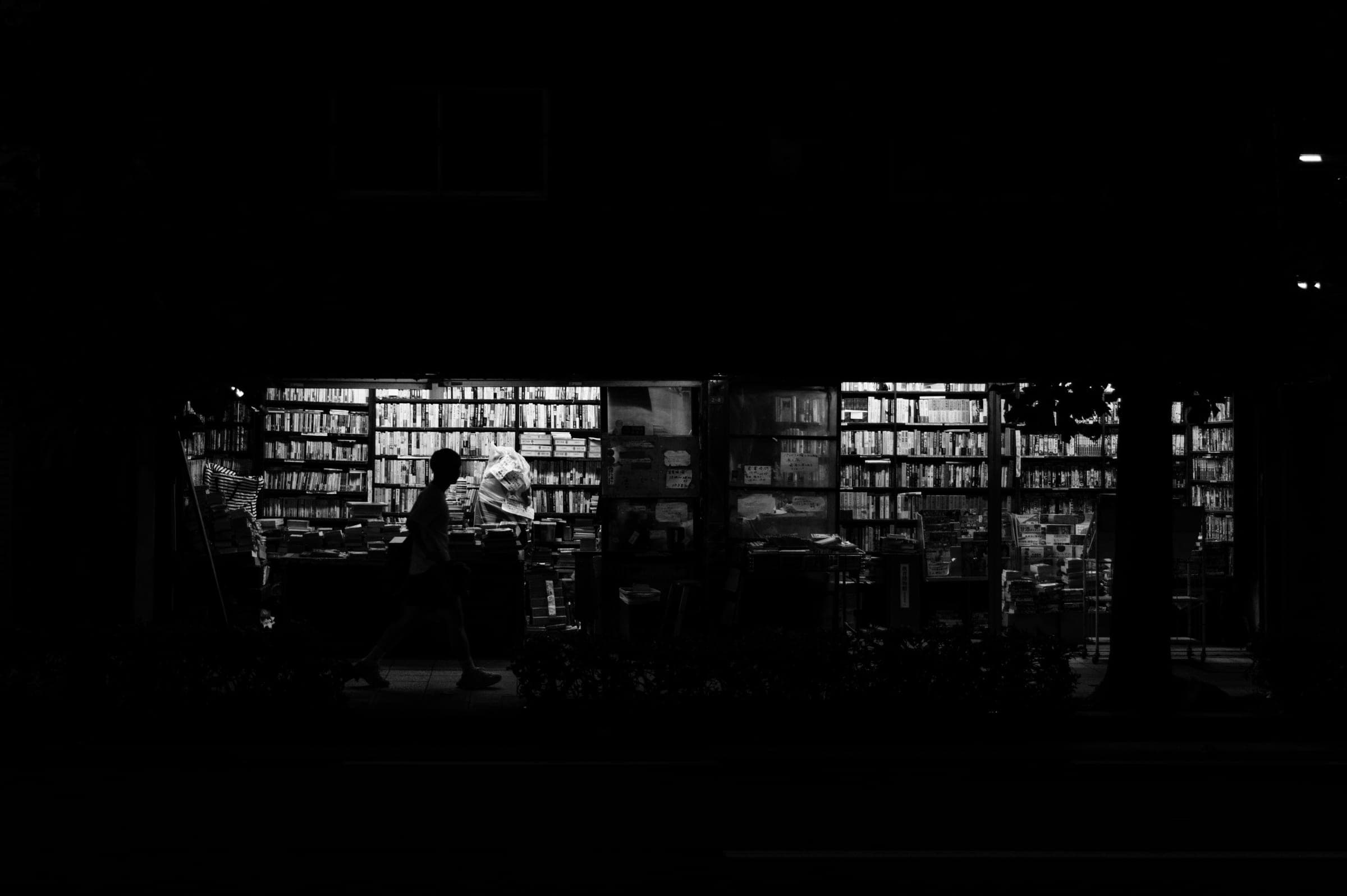 Noir et blanc : un passant marche seul devant une librairie éclairée la nuit, les rayonnages pleins derrière lui, la rue plongée dans le noir.