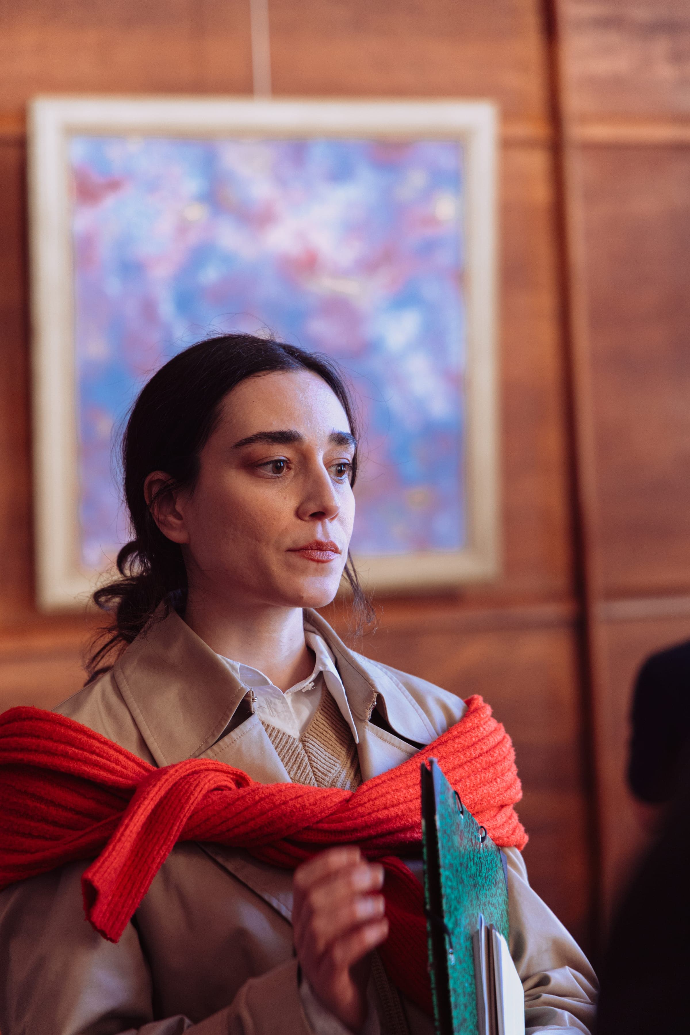 La comédienne en trench et écharpe rouge tient un carnet devant le tableau.