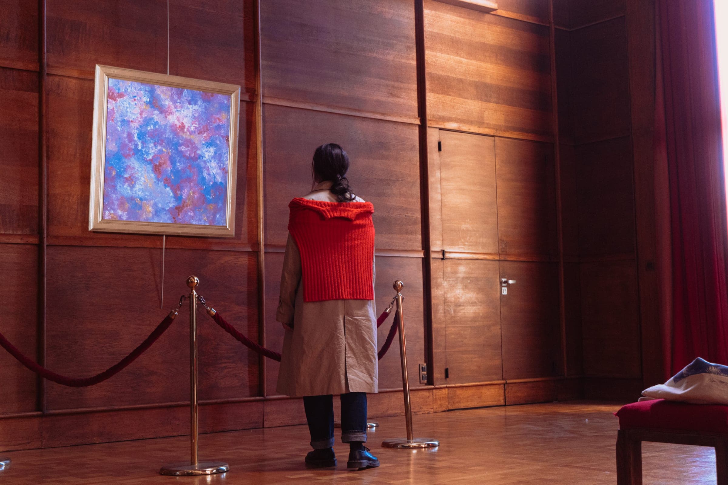 La comédienne à l'écharpe rouge, seule et de dos, devant le tableau derrière le cordon.