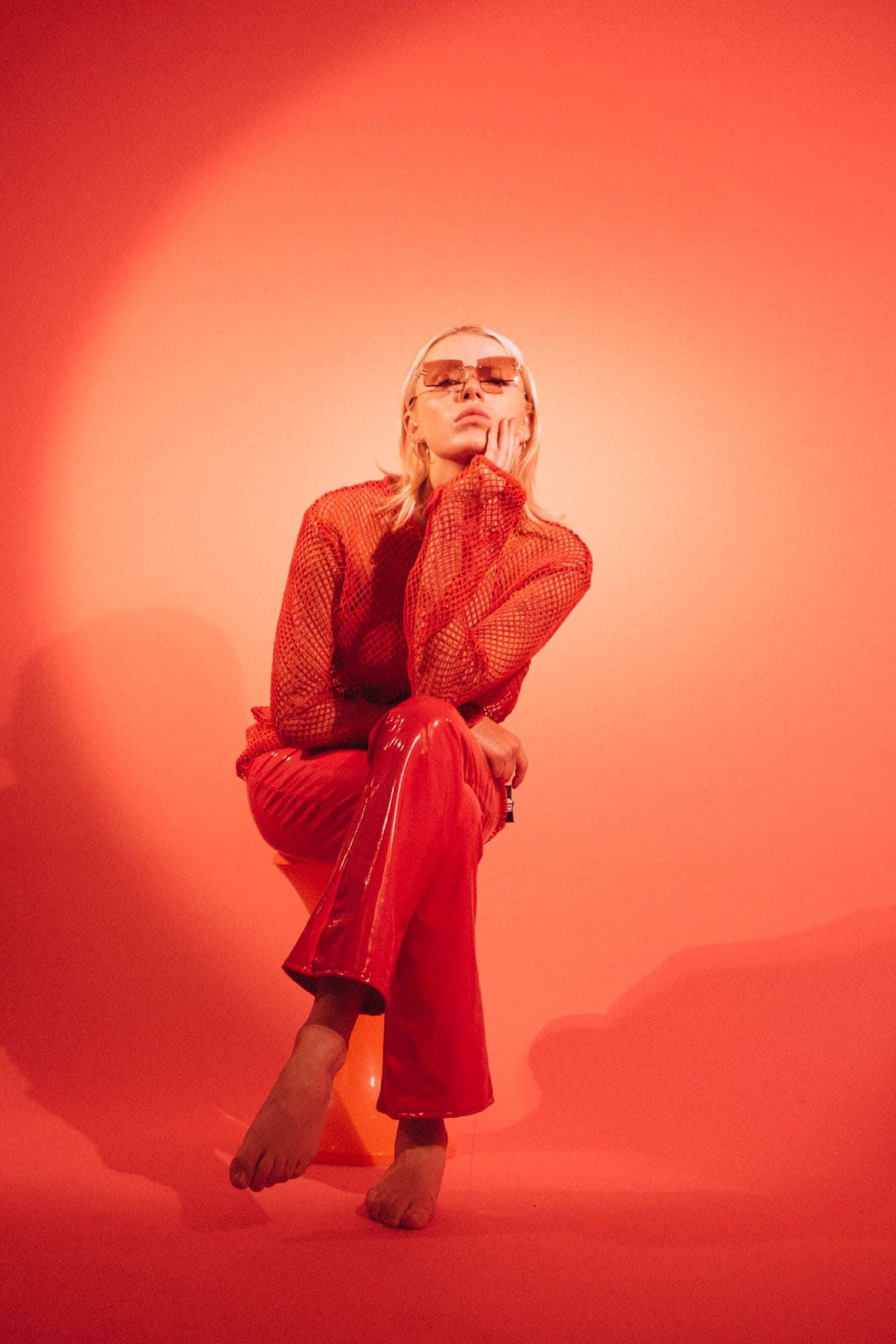 Assise en pied sur un tabouret, résille rouge et pantalon de vinyle rouge évasé, pieds nus, longue ombre portée.
