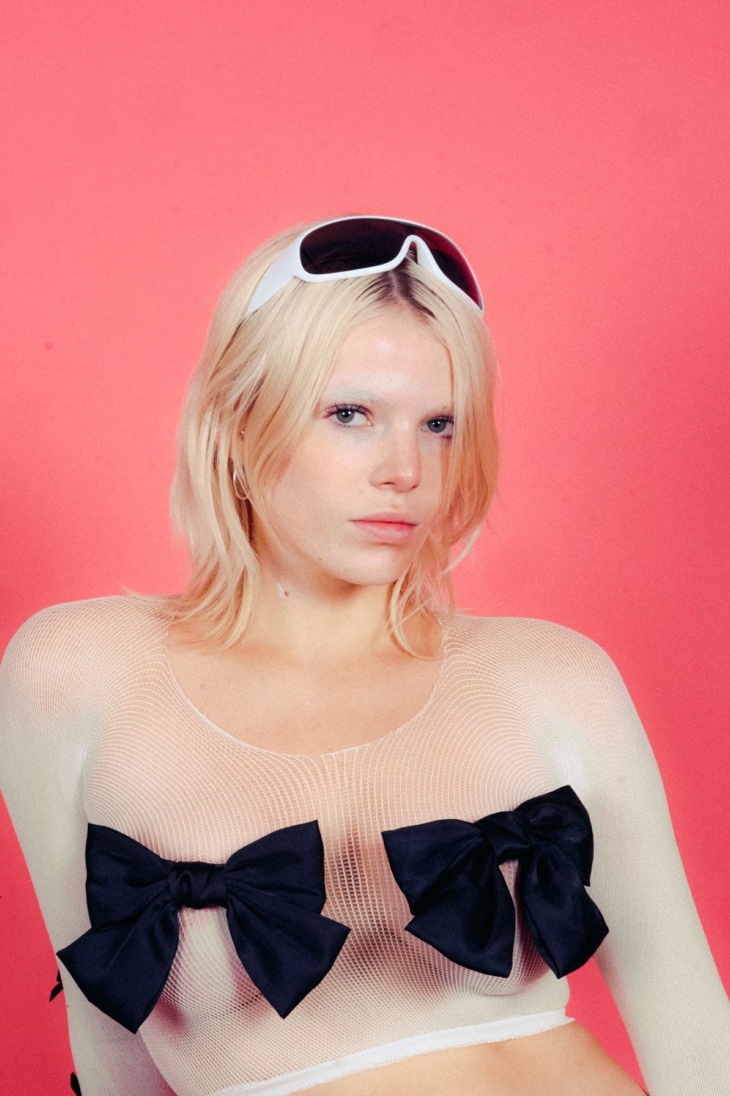 Haut de résille écrue orné de deux nœuds noirs, lunettes masque blanches relevées sur les cheveux, fond rose.