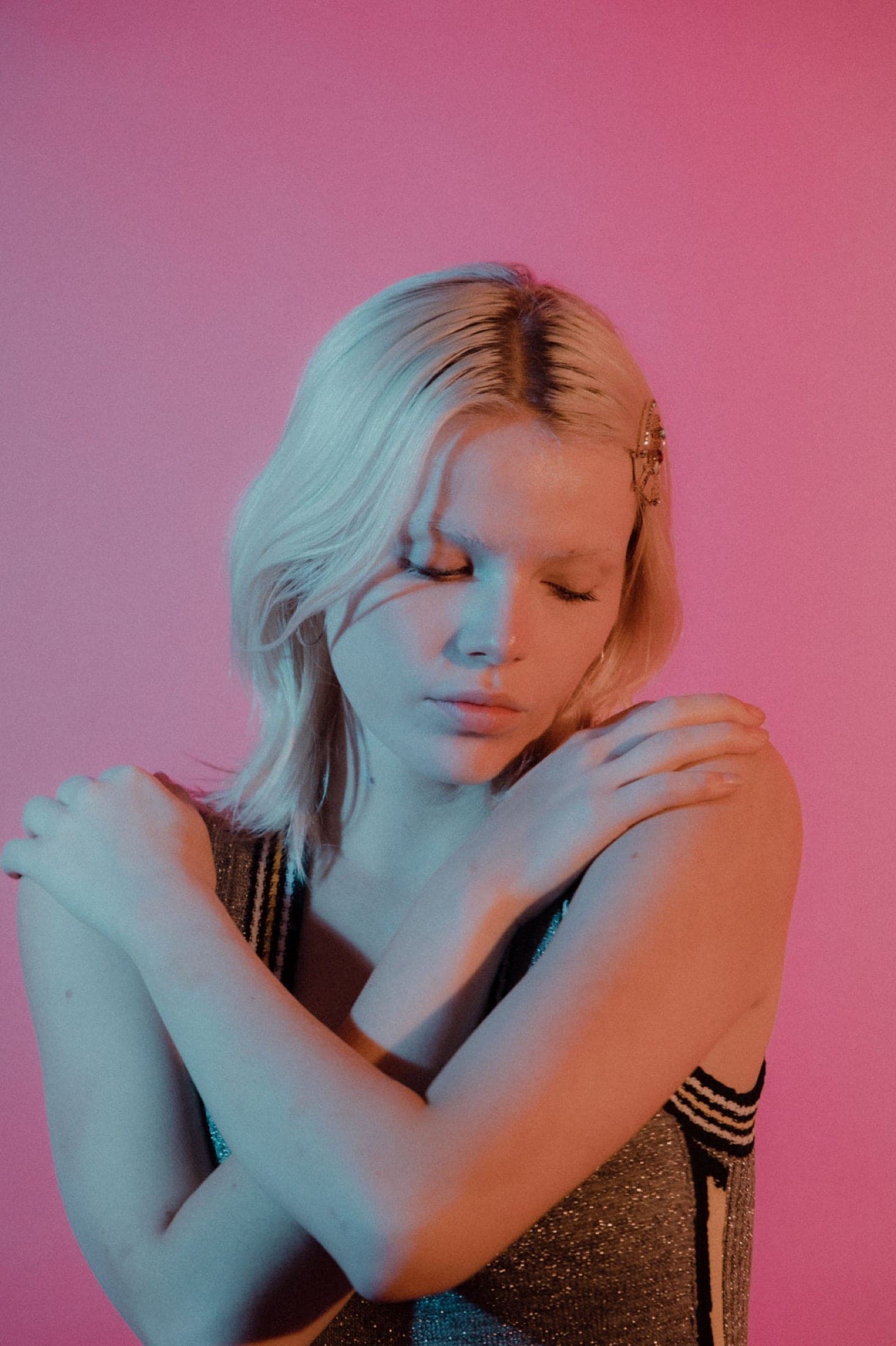 Yeux fermés, bras croisés sur les épaules, débardeur pailleté à rayures, fond rose.