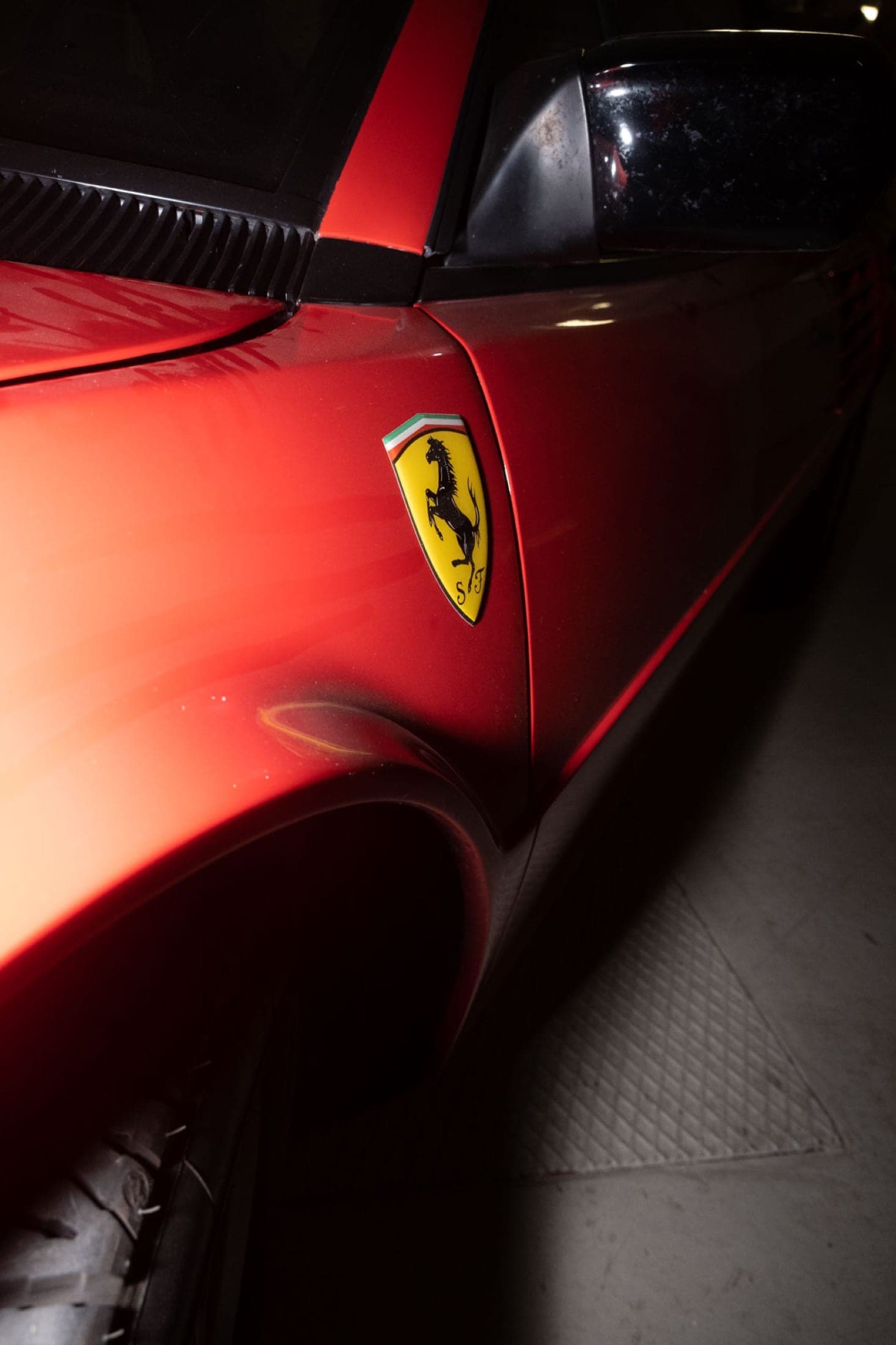 Gros plan sur l'écusson au cheval cabré, sur l'aile rouge d'une Ferrari garée dans un parking sombre.