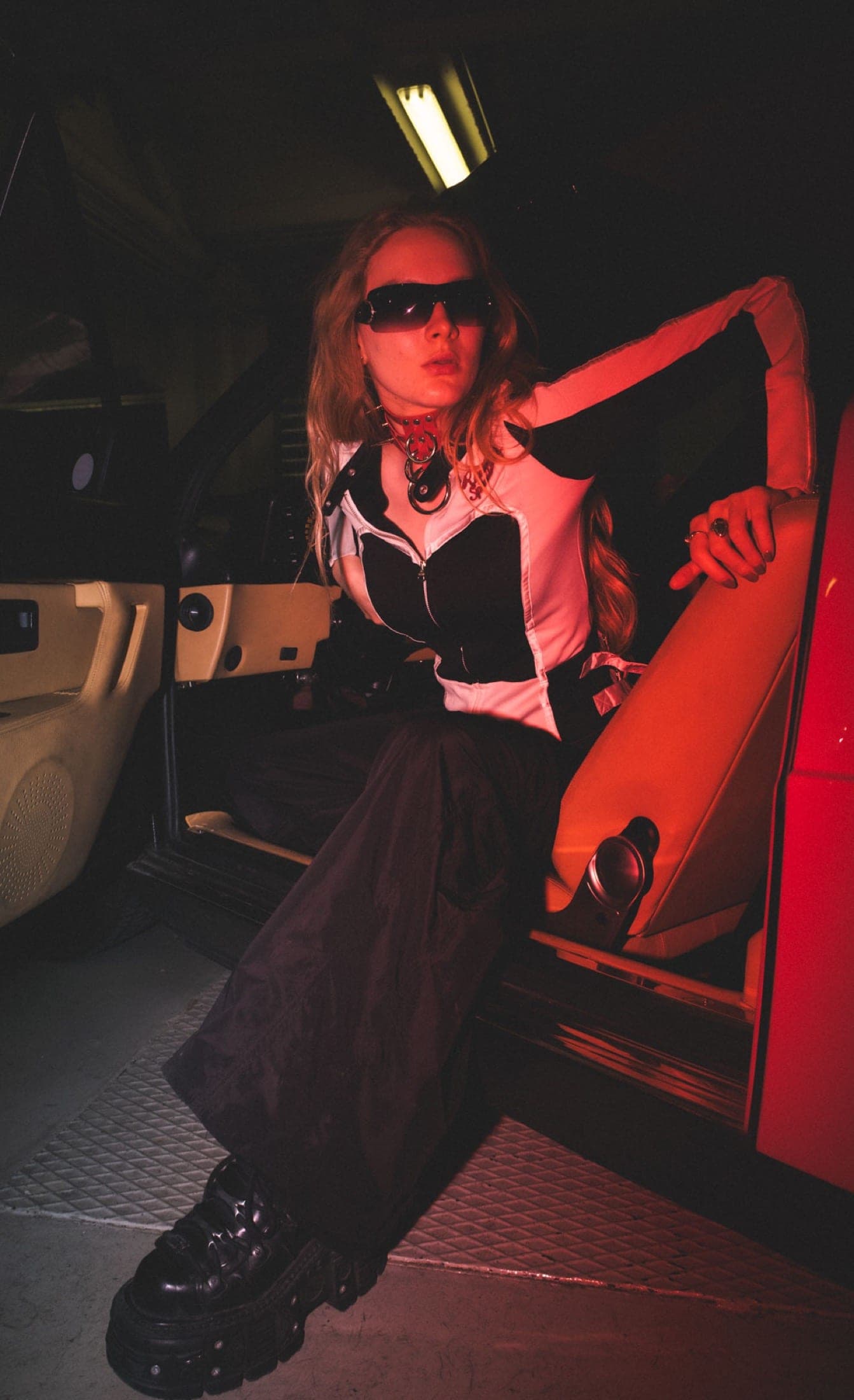 Assis dans l'encadrement de la portière, corset noir et blanc, collier rouge, pantalon large et bottines à plateforme.