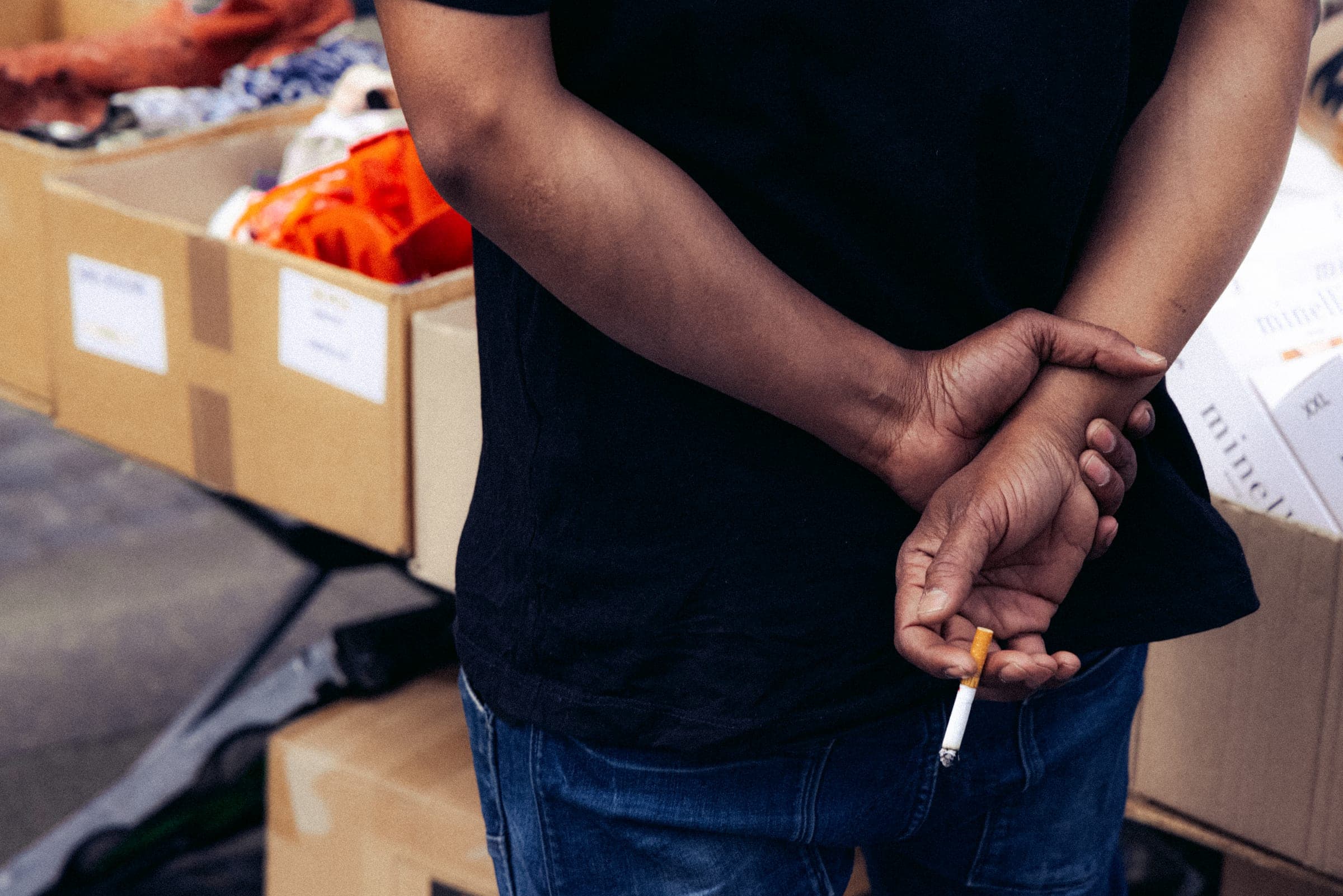Un vendeur, mains croisées dans le dos et cigarette aux doigts, devant ses cageots.