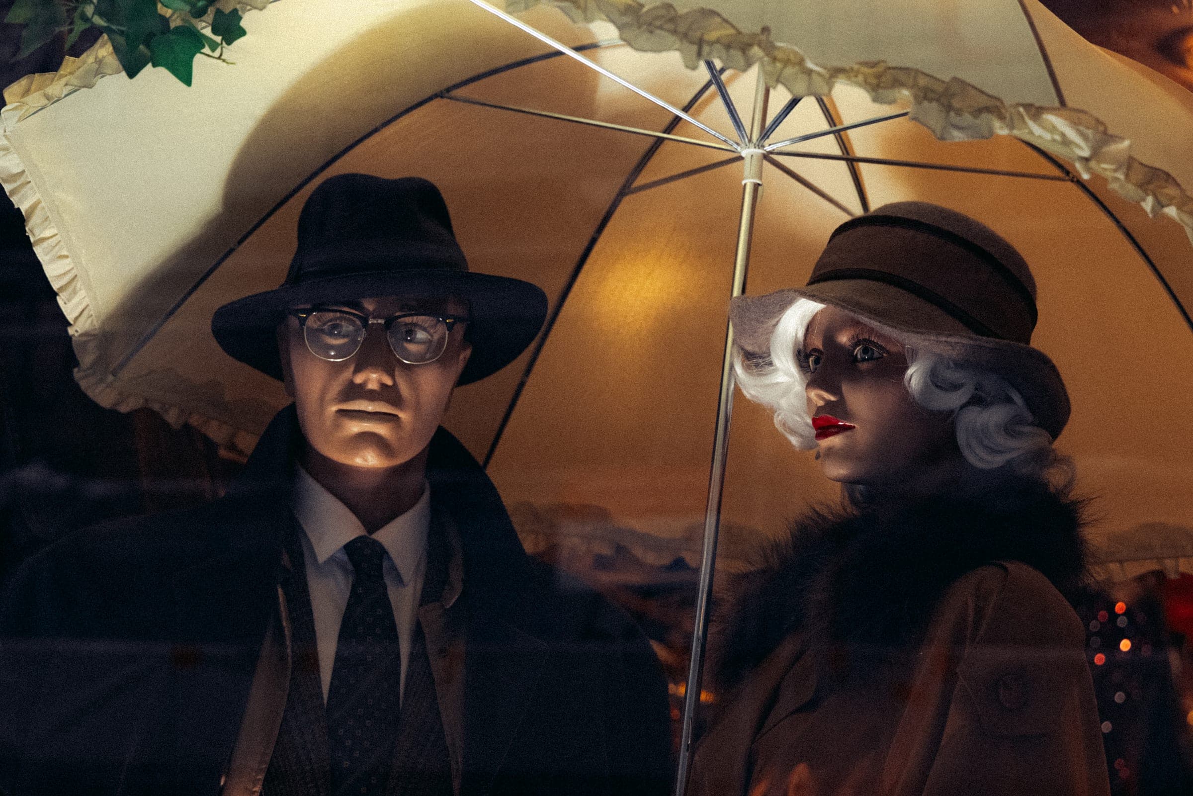 Deux mannequins de vitrine, chapeau et parapluie, dans une lumière dorée.
