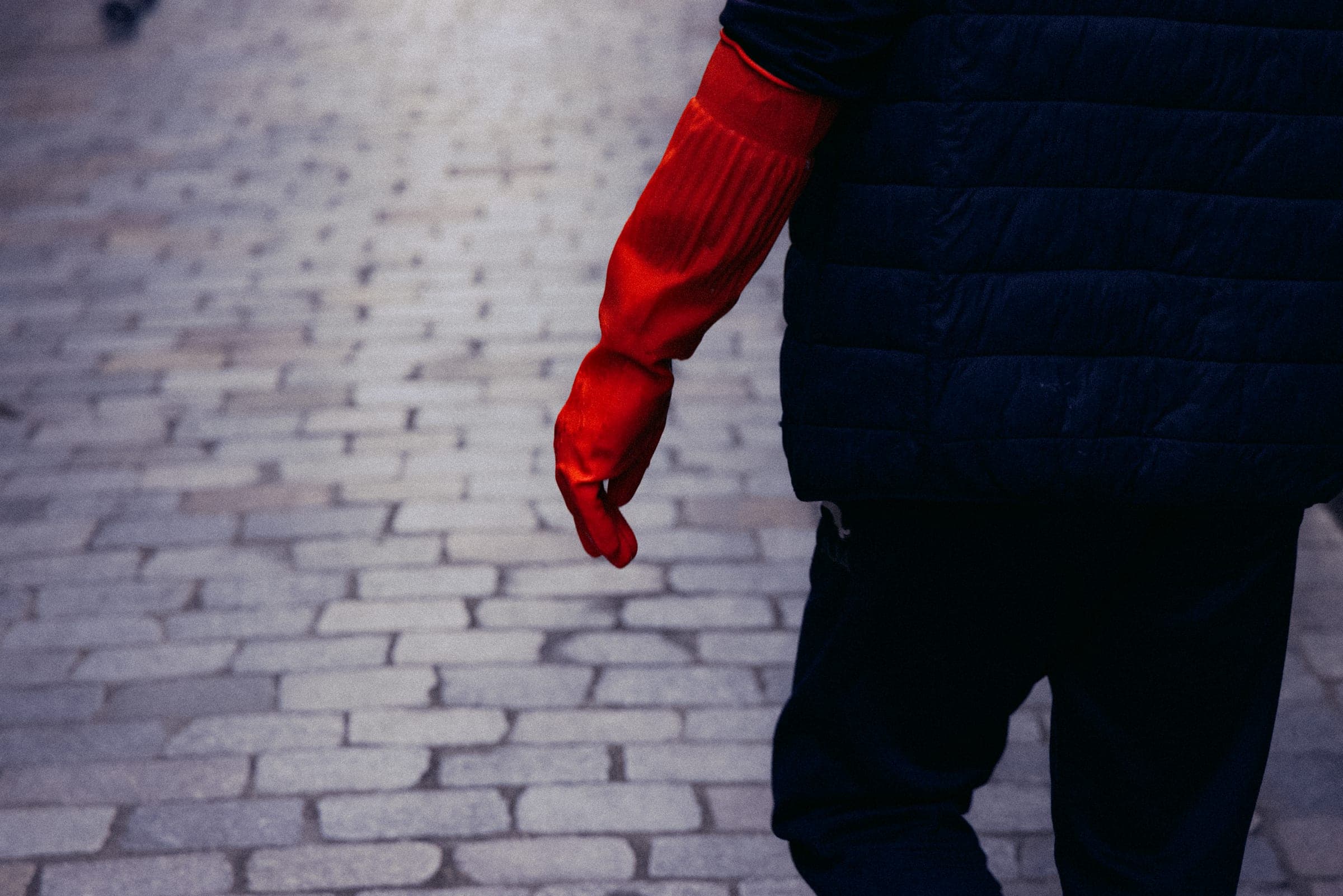 Une main gantée de rouge le long d'un manteau sombre, pavés au sol.