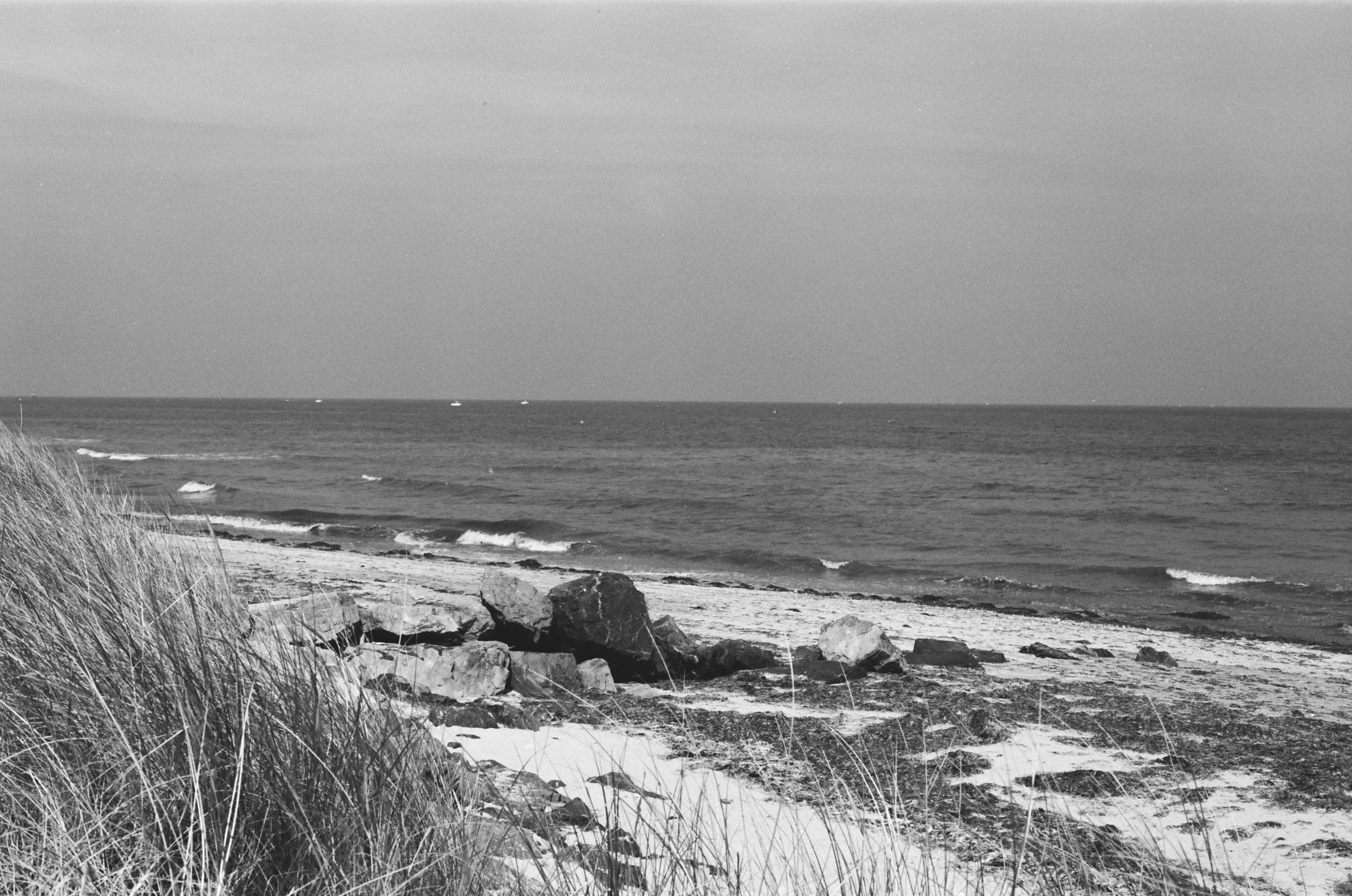 Des oyats au premier plan, quelques rochers sur le sable et la mer sous un ciel gris.